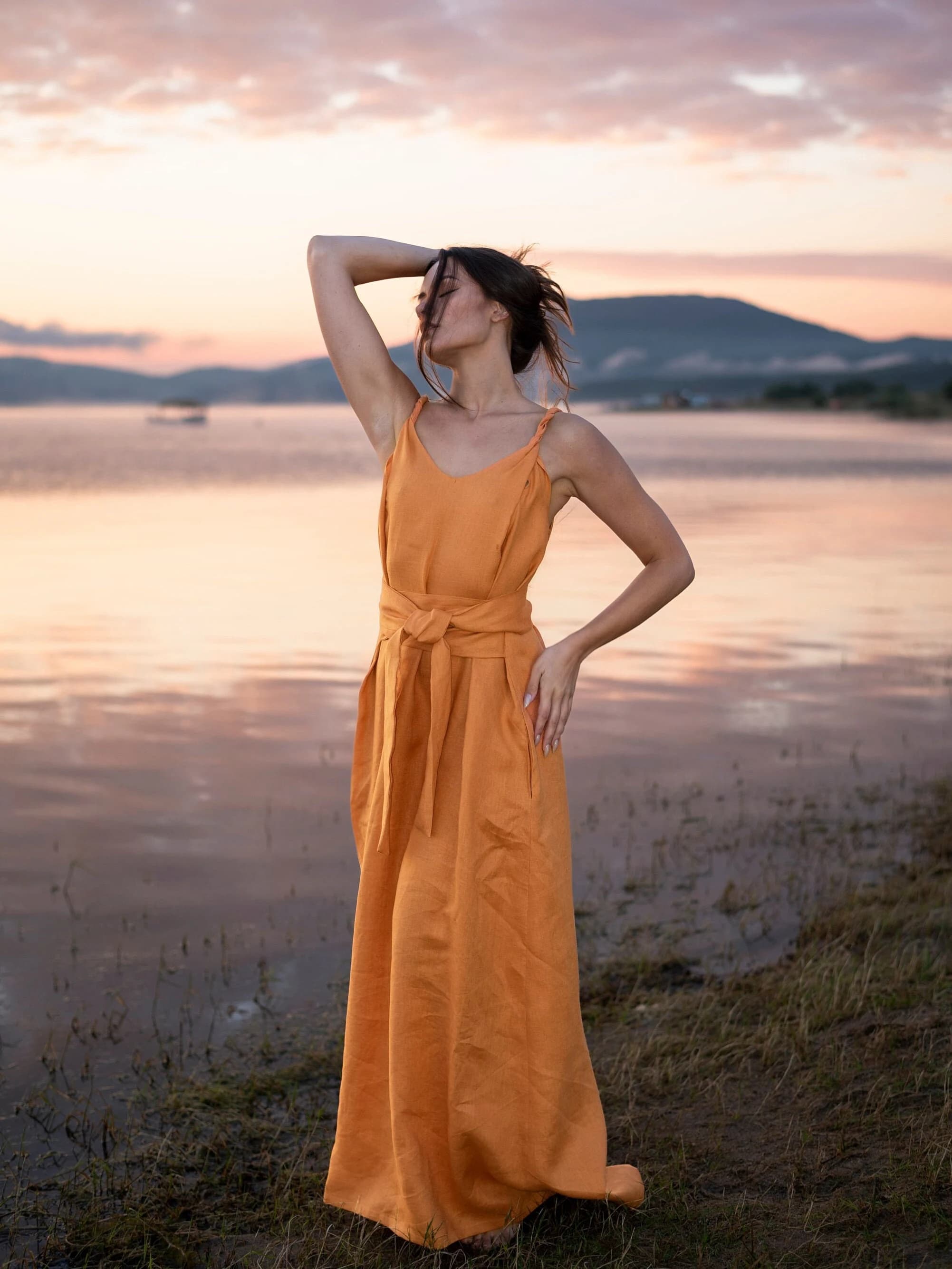 May include: A woman wearing a long, orange linen dress with a tie belt stands on a grassy shore by a lake at sunset.