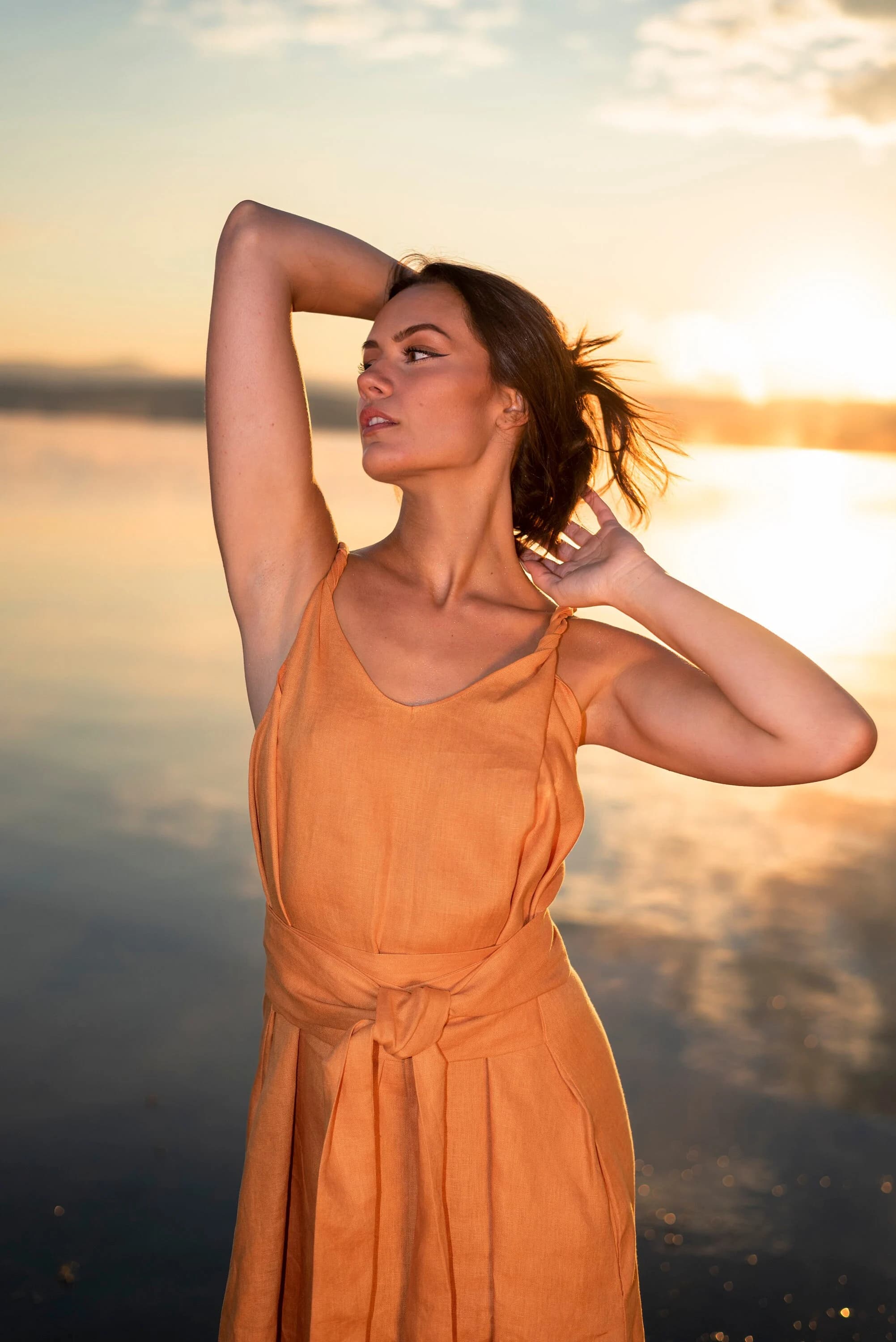 May include: A woman in a loose-fitting, orange linen dress with a tie belt stands in front of a body of water at sunset. The dress has a V-neckline and a relaxed fit.