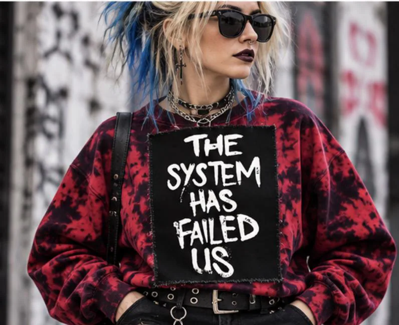 Blonde woman with blue hair wearing a red and black tie-dye sweatshirt with The System Has Failed Us punk patch sewn on the front.