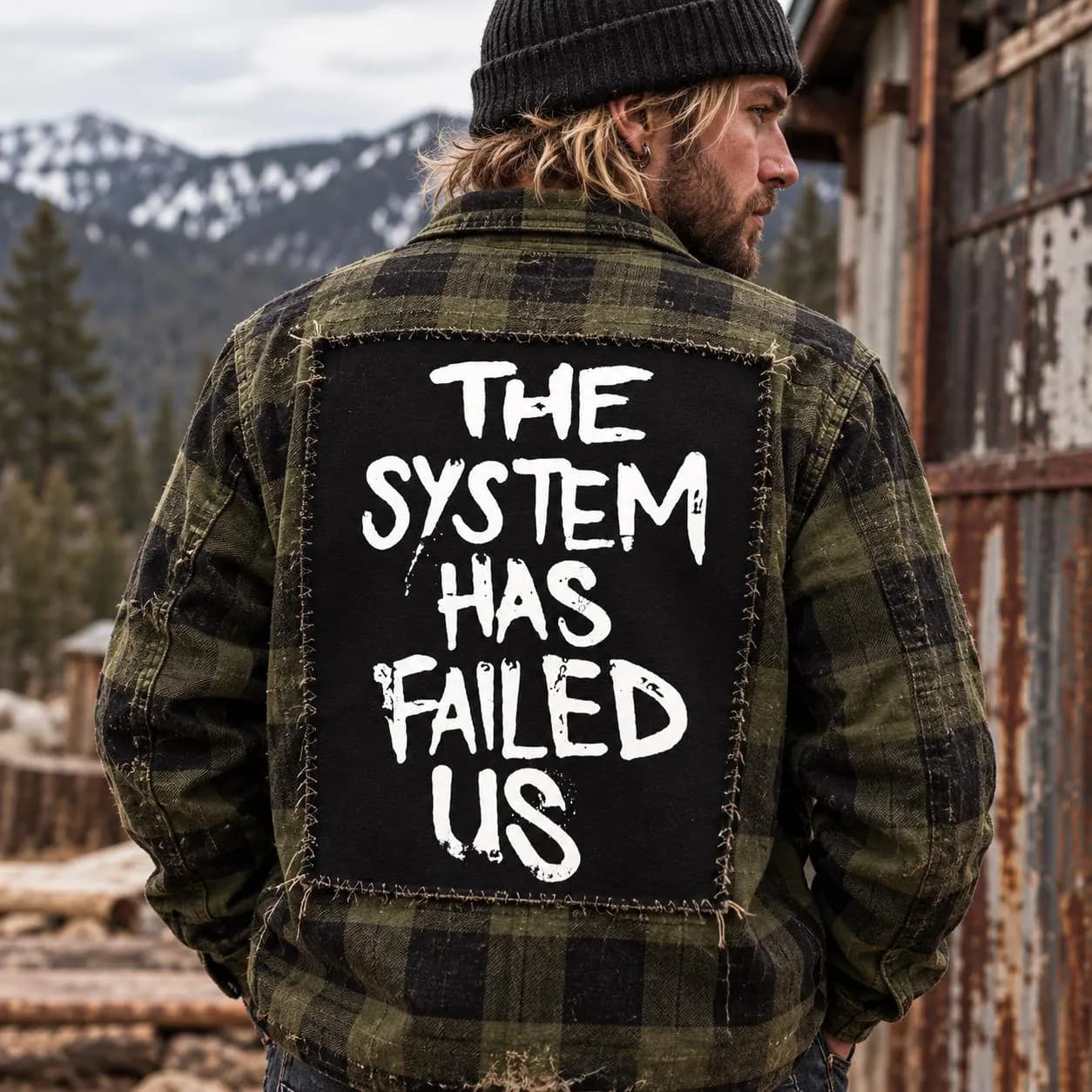 Male punk model wearing a rustic green plaid flannel jacket and beanie with large The System Has Failed Us back patch in a mountain setting.