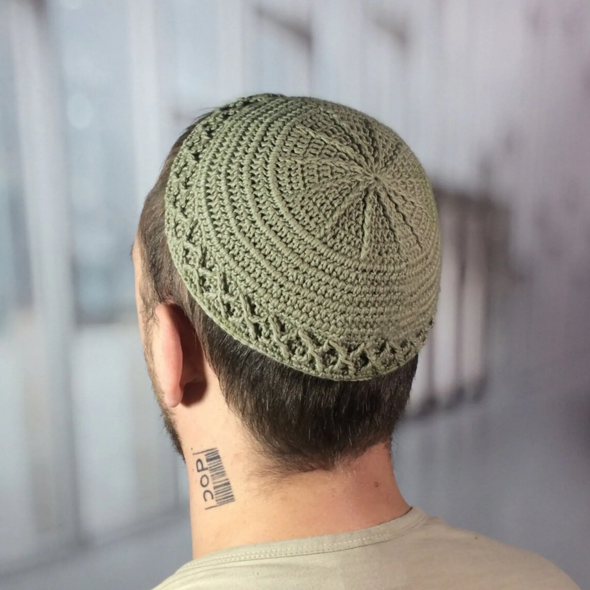 A sage green crocheted kippah, featuring a circular design with a decorative border. The handmade head covering is shown from the back, with the wearer's dark hair visible. The kippah is a traditional Jewish head covering.