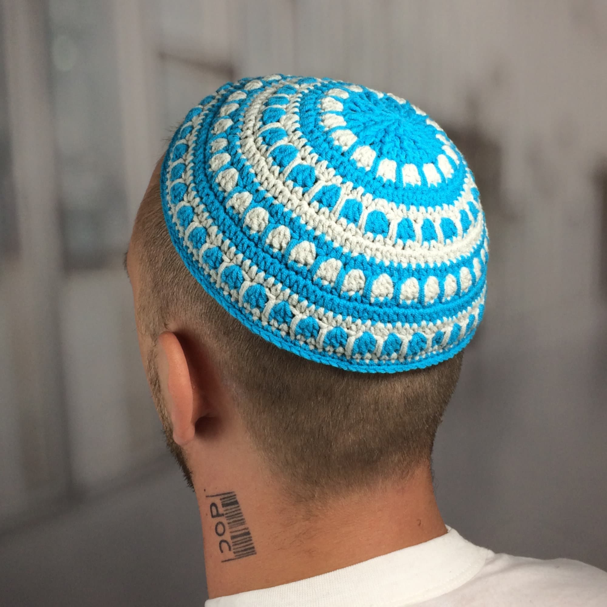 A crocheted kippah in a vibrant blue and white color scheme. The circular cap features a repeating pattern of white and blue crochet stitches. The kippah is worn on the head.
