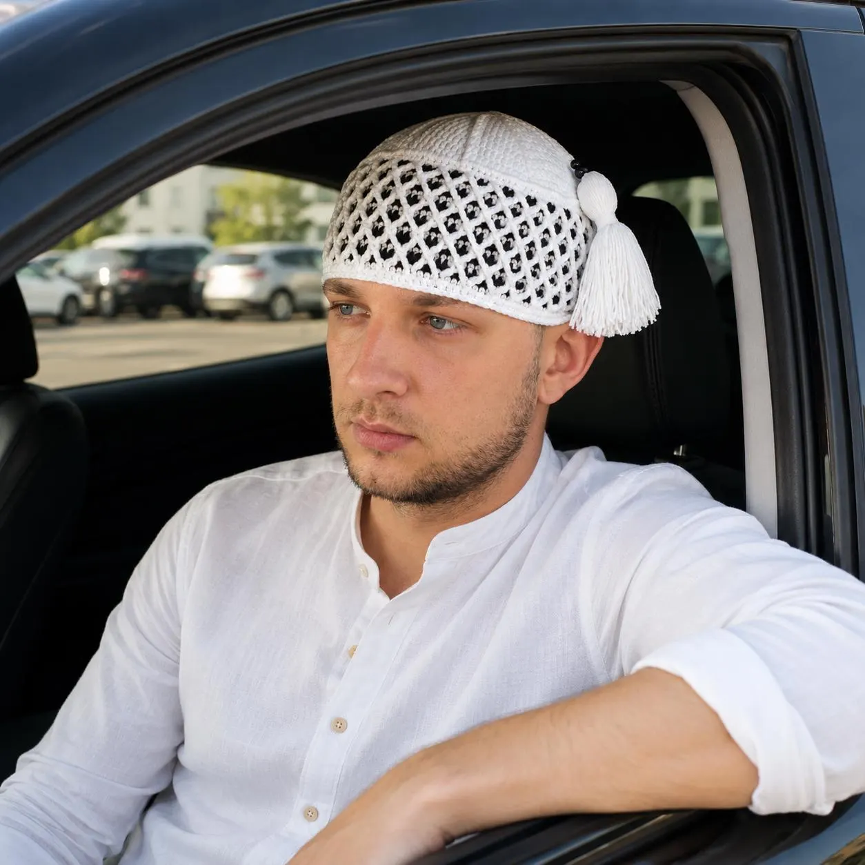 Lifestyle photo of a man sitting inside a car wearing a clean white shirt and a white crochet kufi cap with a matching white tassel.