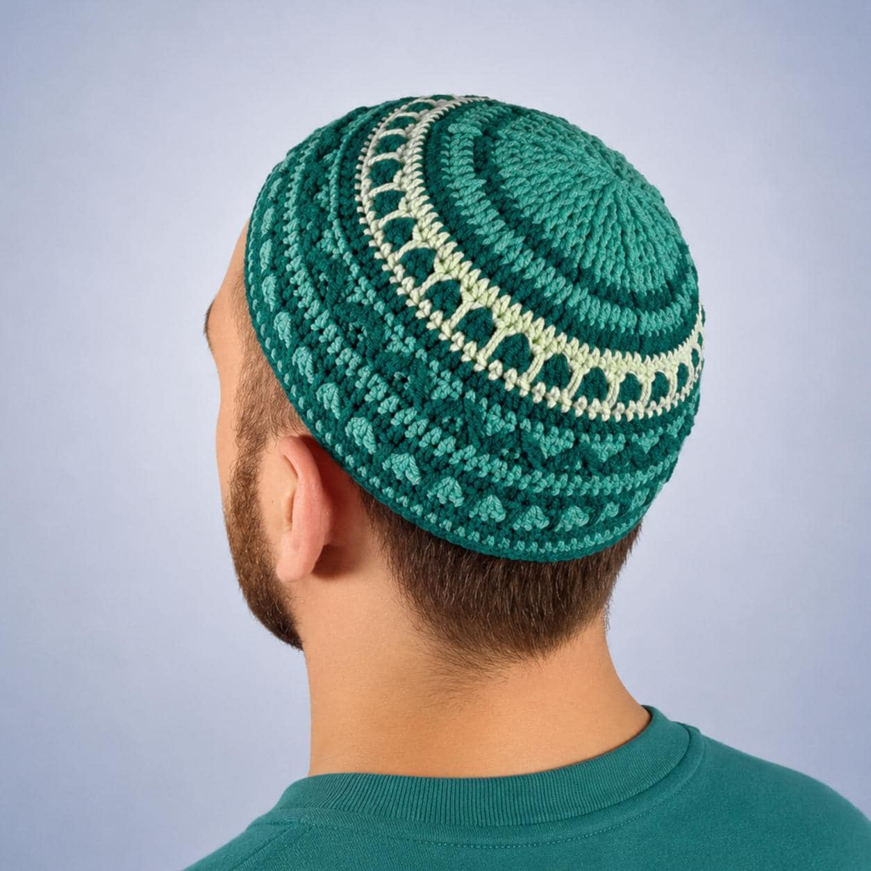 A handmade crochet hat in shades of teal and light yellow. The kippah features a textured pattern with a circular design around the crown.