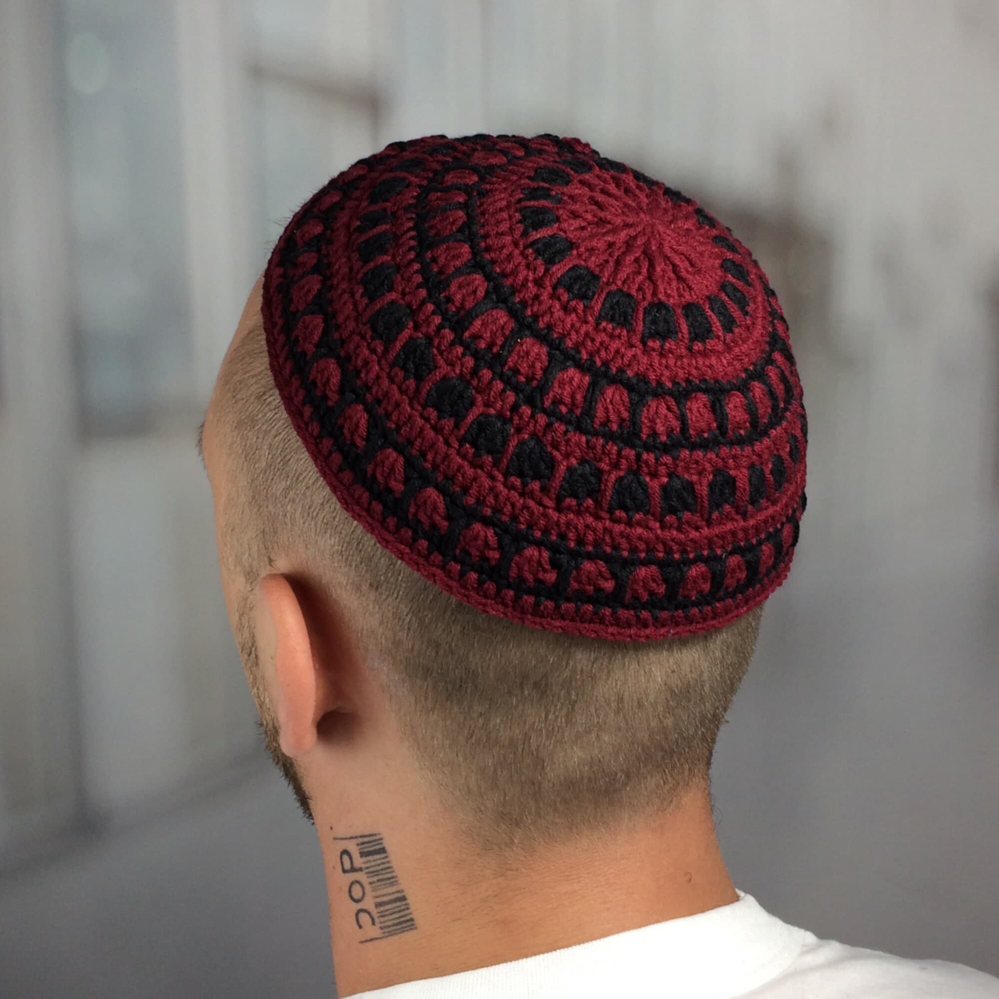 A burgundy and black crocheted kippah, a traditional Jewish head covering. The kippah features a circular design with alternating bands of burgundy and black. The head covering is worn by a person with short, light-colored hair.