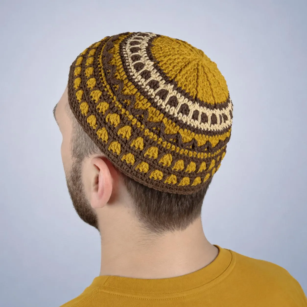 A crocheted skull cap in shades of mustard yellow, brown, and cream. The kippah features a detailed pattern with geometric and floral elements. The cap is worn by a person.
