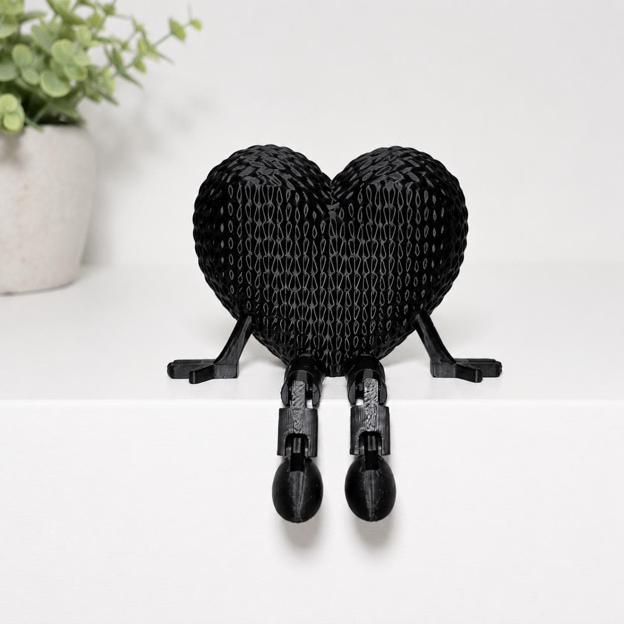 Black articulated heart figurine sitting on a white shelf with dangling legs, photographed in a clean minimalist aesthetic background for Etsy listing.