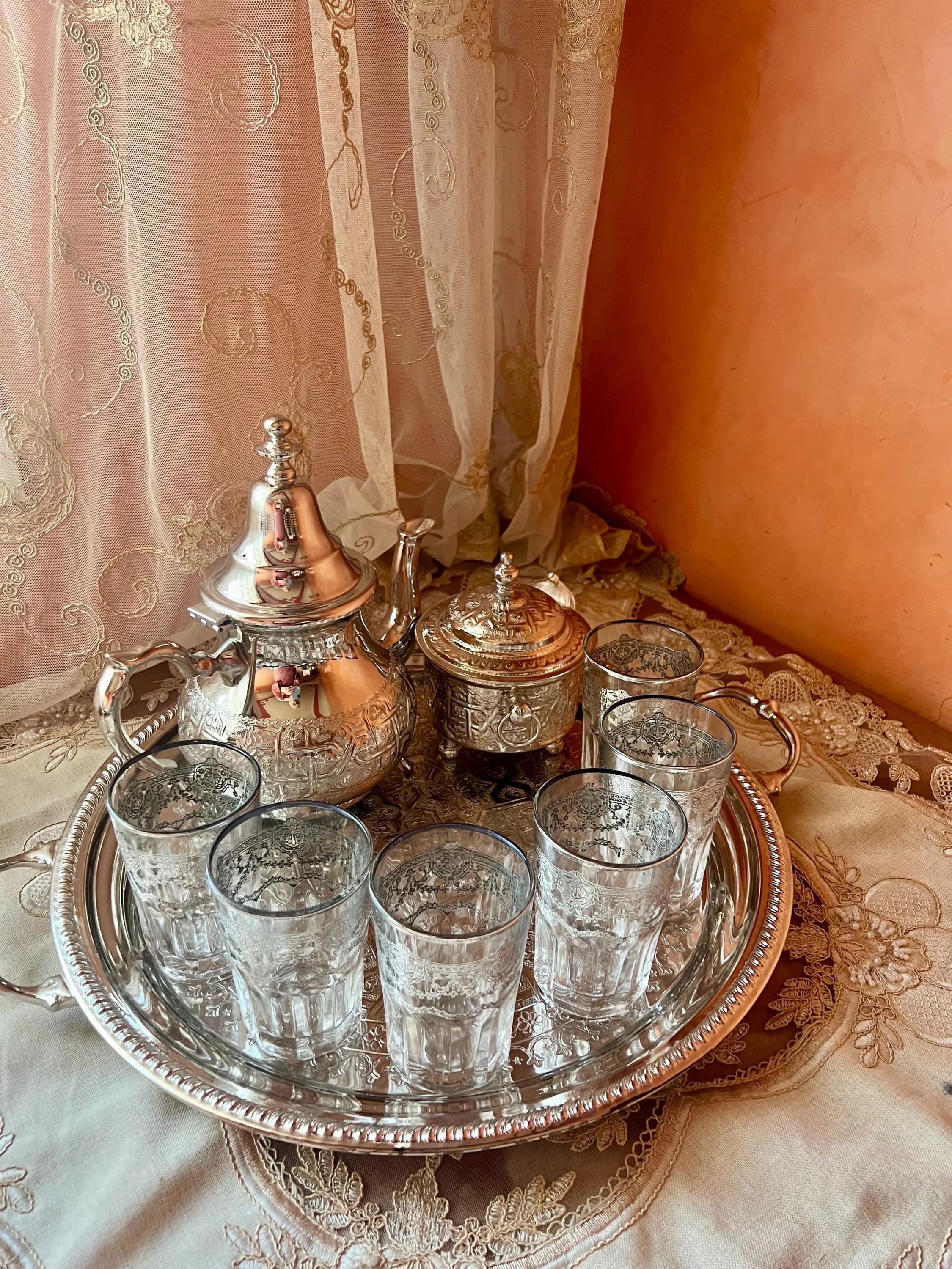 A close-up of a silver Moroccan tea set on a lace tablecloth, displaying an engraved teapot, sugar bowl, and six glasses.