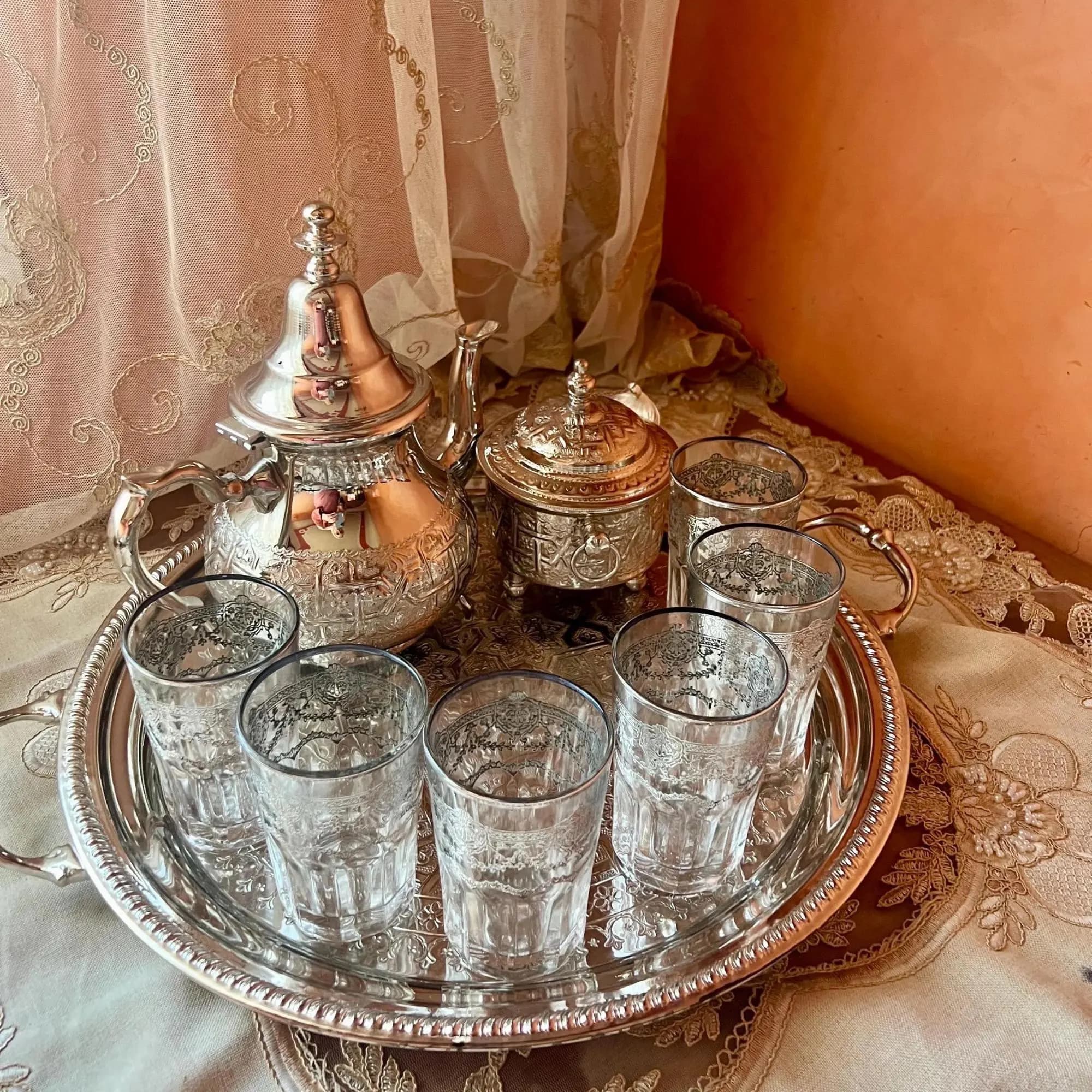 A silver Moroccan tea set set up on a decorative tray over a patterned fabric surface.