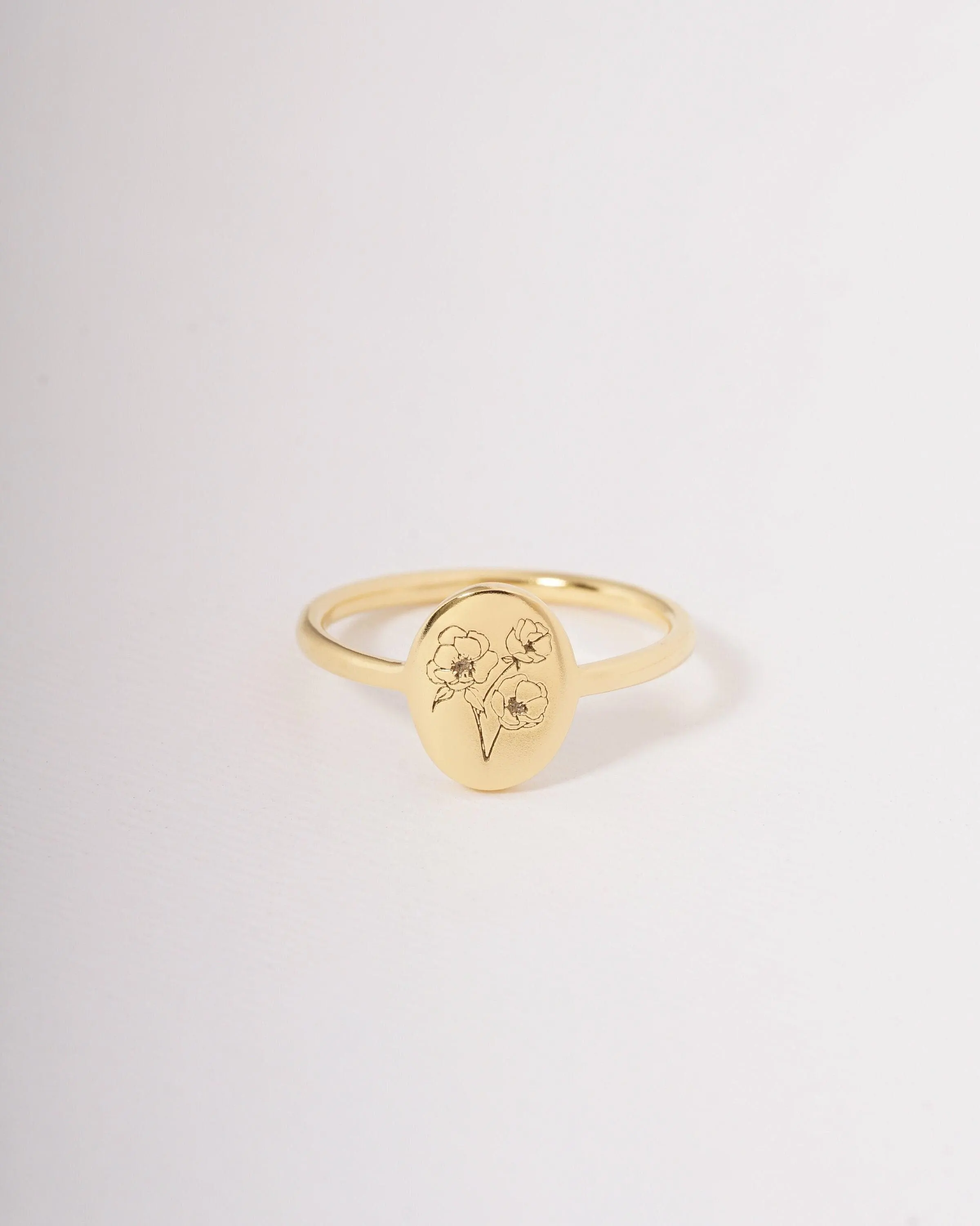 May include: A gold-colored ring with an oval face featuring a floral design. The ring has a thin band and a smooth, polished finish. The floral design is a delicate, engraved illustration of three flowers.