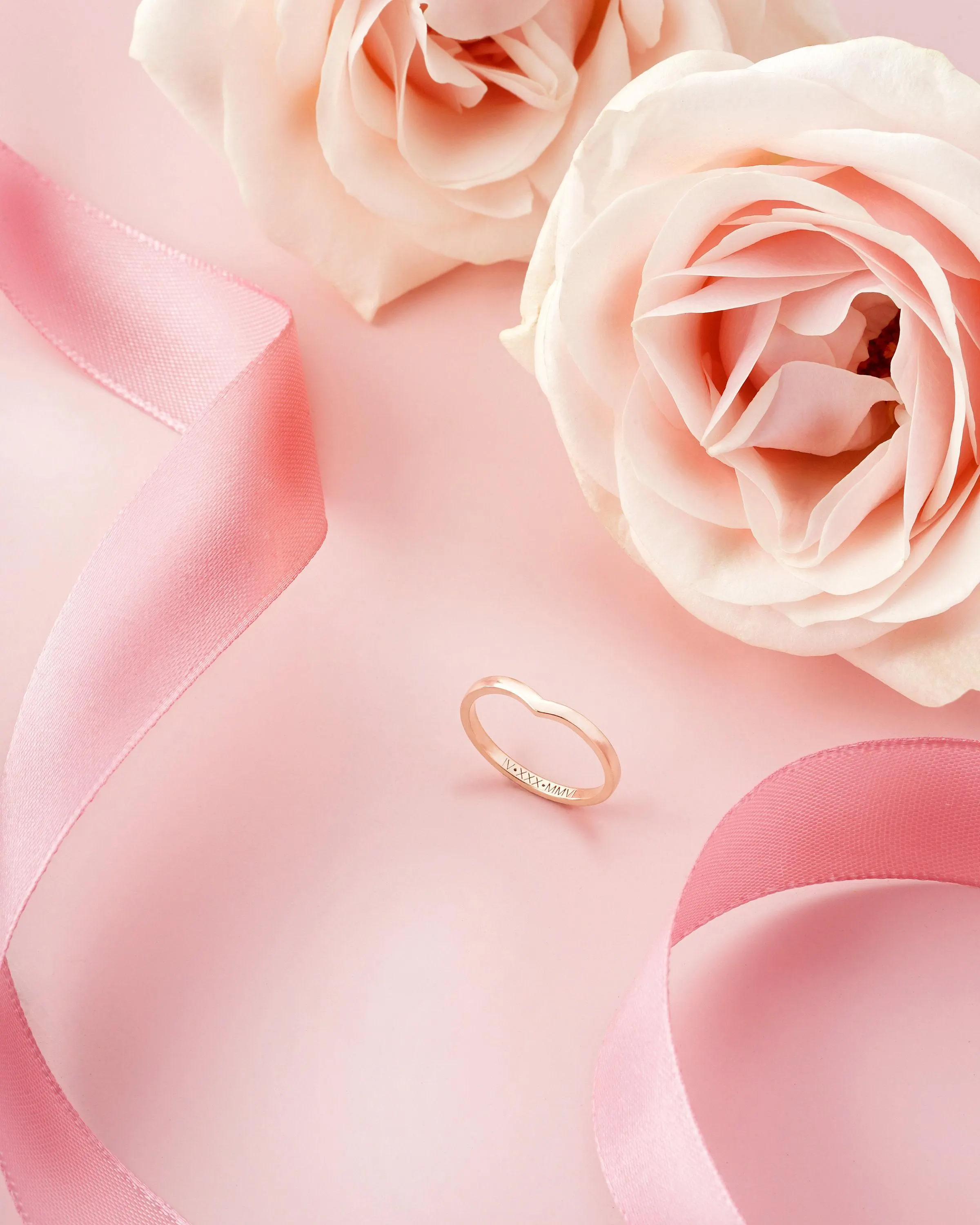 May include: A gold-colored ring with a V-shaped design, resting on a pink surface. Two light pink roses and a pink ribbon are also present. The ring has text engraved on it.