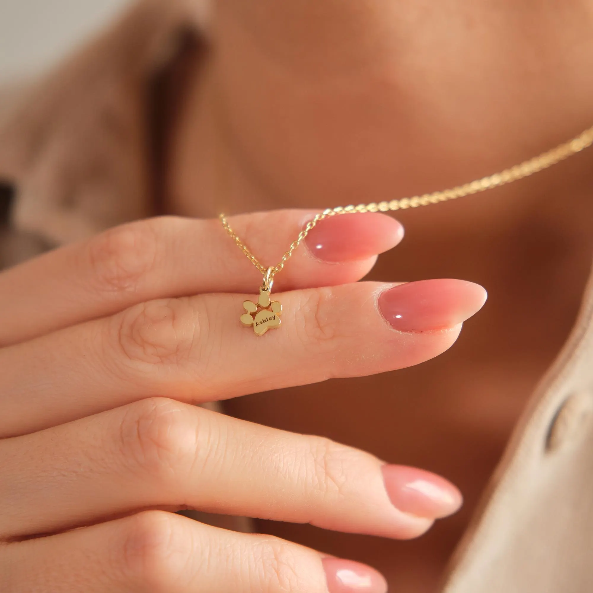 “14k solid gold paw print name necklace held between fingers, personalized pet jewelry charm.”