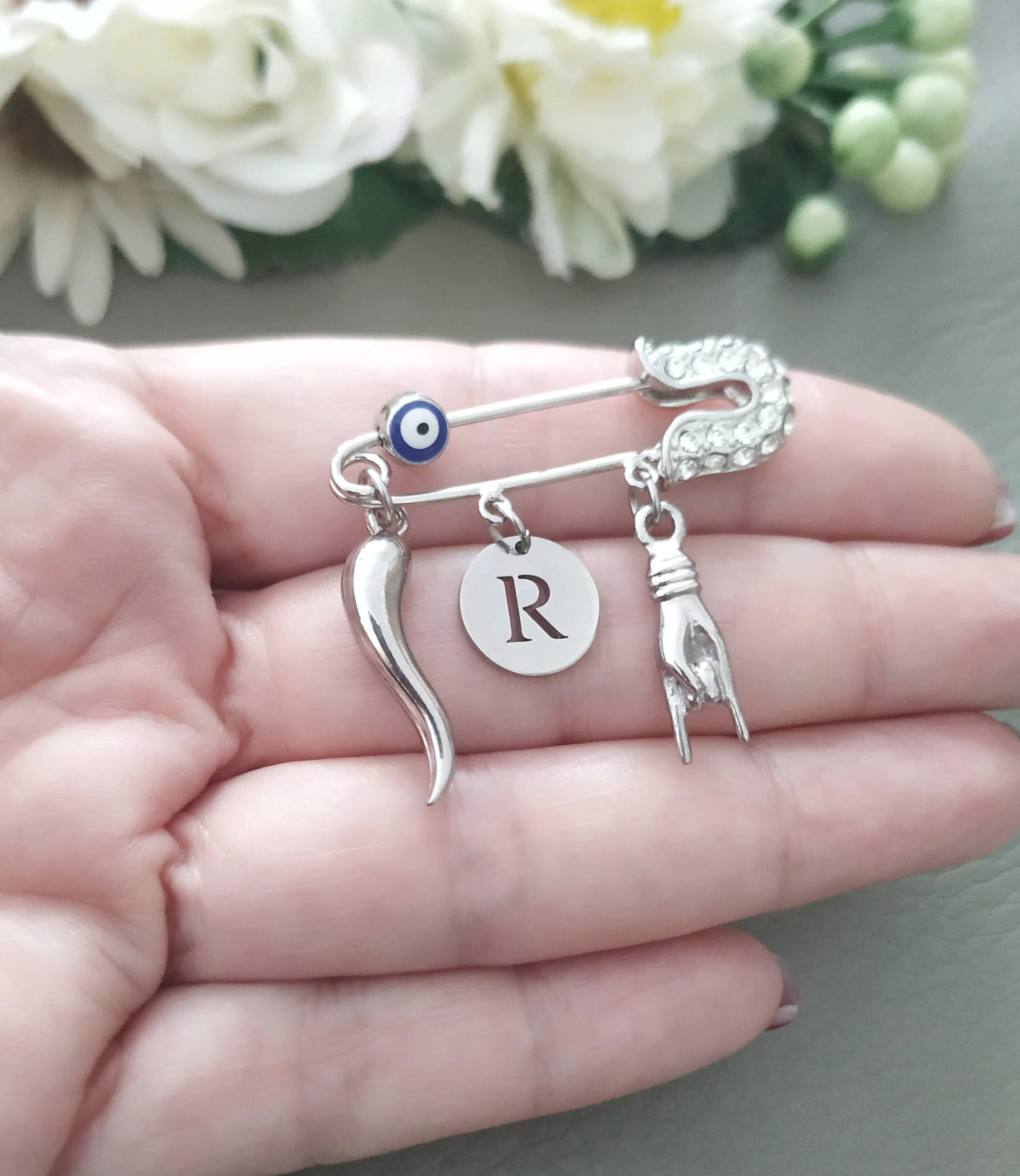 A silver-tone safety pin brooch featuring a small blue evil eye bead, Italian horn charm, a round disc with the letter R, and a mano cornuto hand gesture charm. The safety pin is embellished with clear stones.