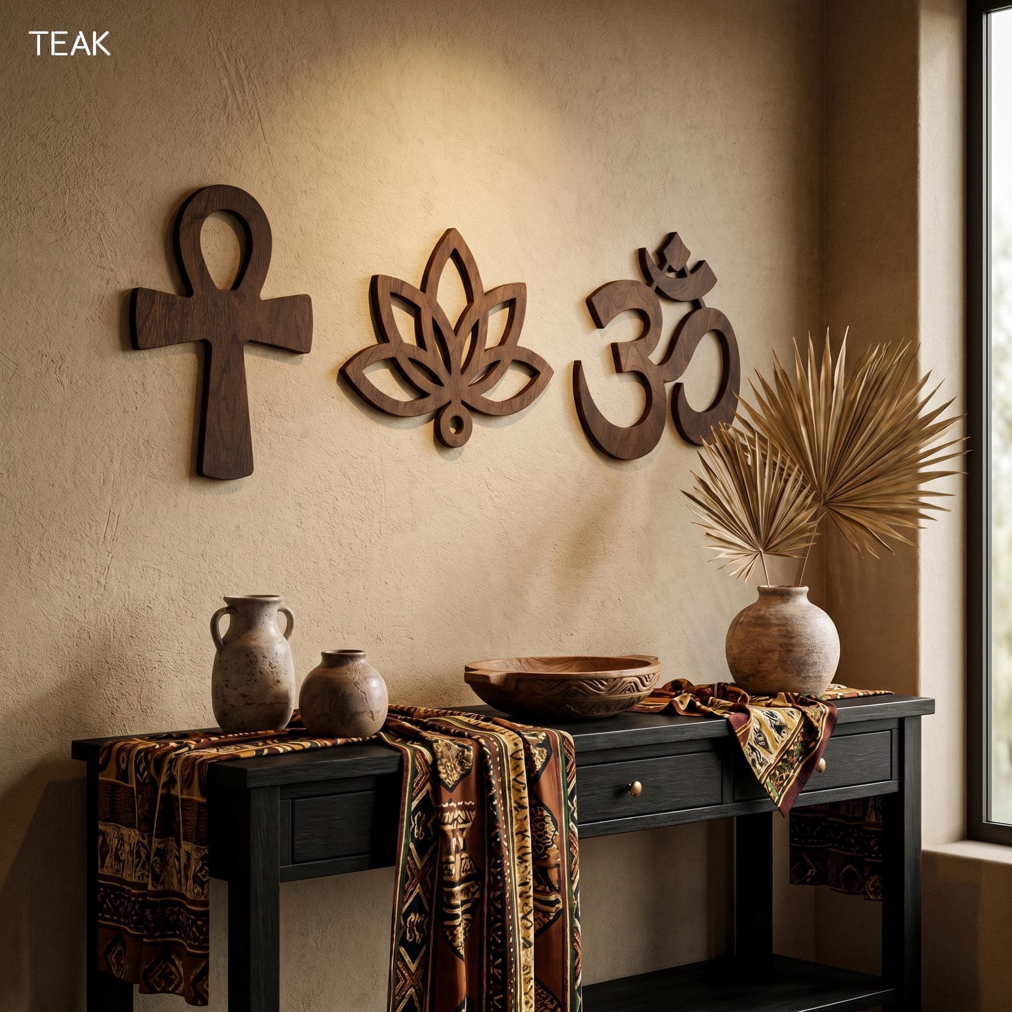 Handmade wooden spiritual wall art set in a teak-style finish mounted on a textured beige wall above a black console table. The display includes an Ankh symbol, lotus flower wall art, and Om wall decor made from cut plywood with visible wood grain and smooth openwork details. Warm sunlight, ceramic vases, patterned textiles, dried palm leaves, and earthy yoga room decor create a calm meditation-inspired interior.