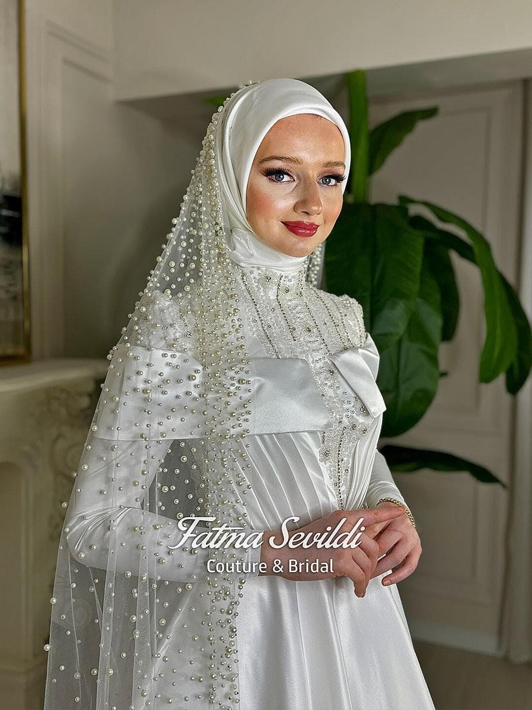 Luxury hijab wedding dress in satin A-line silhouette with high neck, long sleeves, and pearl-beaded tulle veil styling for a modest couture bridal look. Crafted for elegant Muslim and modest brides.