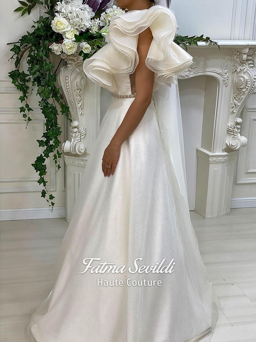 Sparkly A-line wedding dress with dramatic ruffle sleeves, illusion V-neckline, and a beaded belt in ivory couture style. Lightweight glitter tulle finish for a modern bridal look.