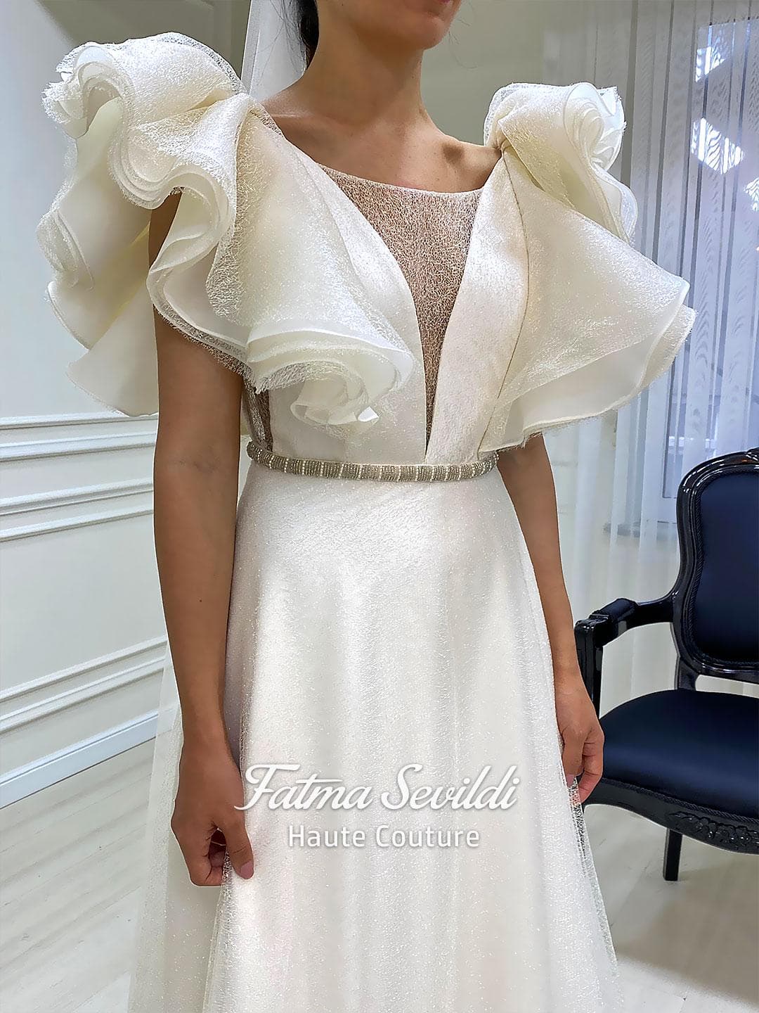 Sparkly A-line wedding dress with dramatic ruffle sleeves, illusion V-neckline, and a beaded belt in ivory couture style. Lightweight glitter tulle finish for a modern bridal look.