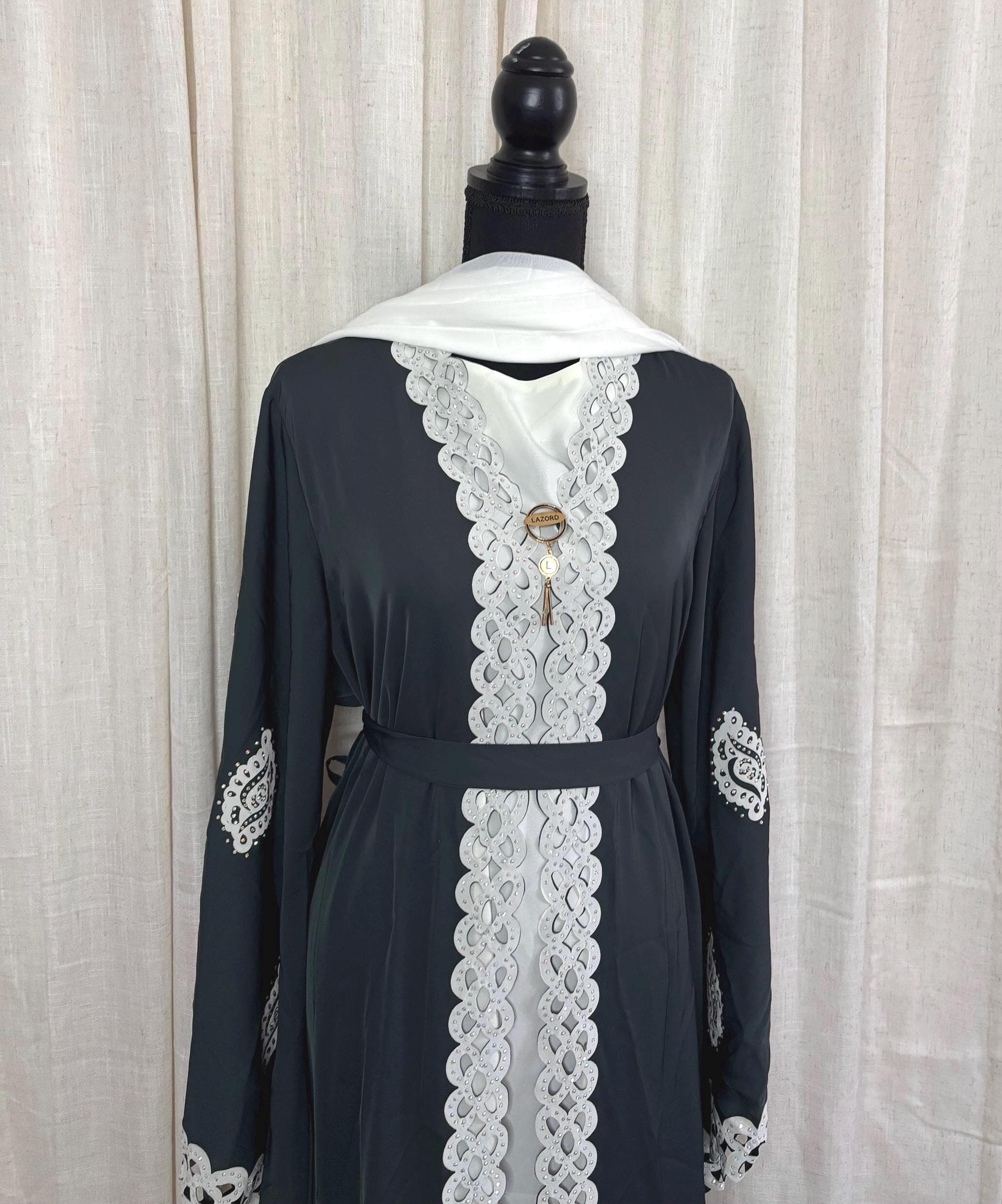 May include: A black abaya with white lace trim and a white scarf. The abaya has a key-shaped pendant hanging from the neckline.