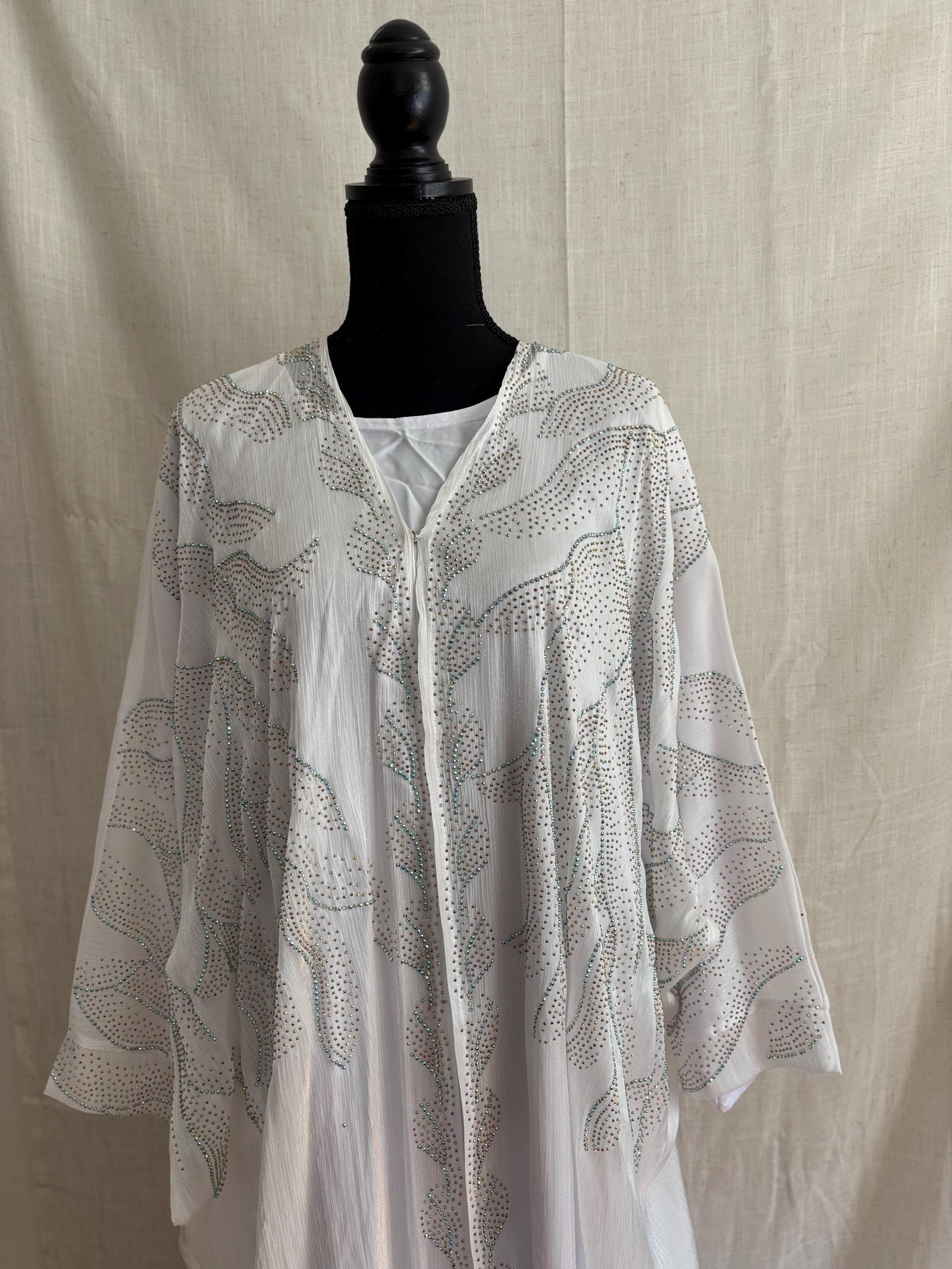 May include: White, flowing garment with silver-colored embellishments. The long-sleeved top features a V-neck and a pattern of dotted lines. The garment is displayed on a black mannequin head.