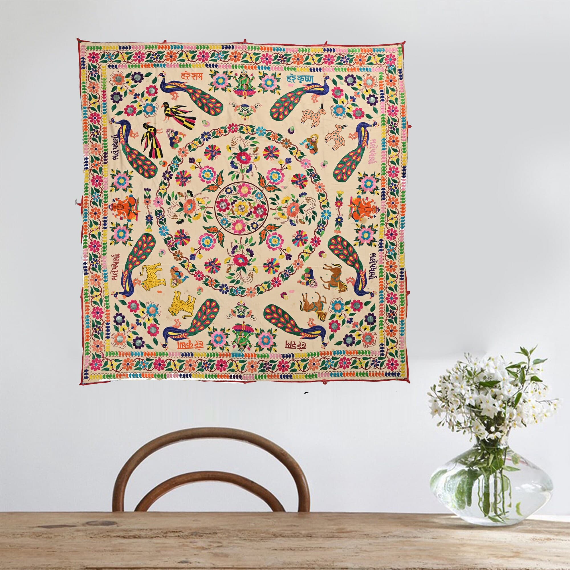 May include: Wall hanging Boho Banjara square, cream-colored tapestry with a colorful, circular design. The design features peacocks, flowers, animals, and text in a Hindi language. The tapestry is surrounded by a colorful border.
