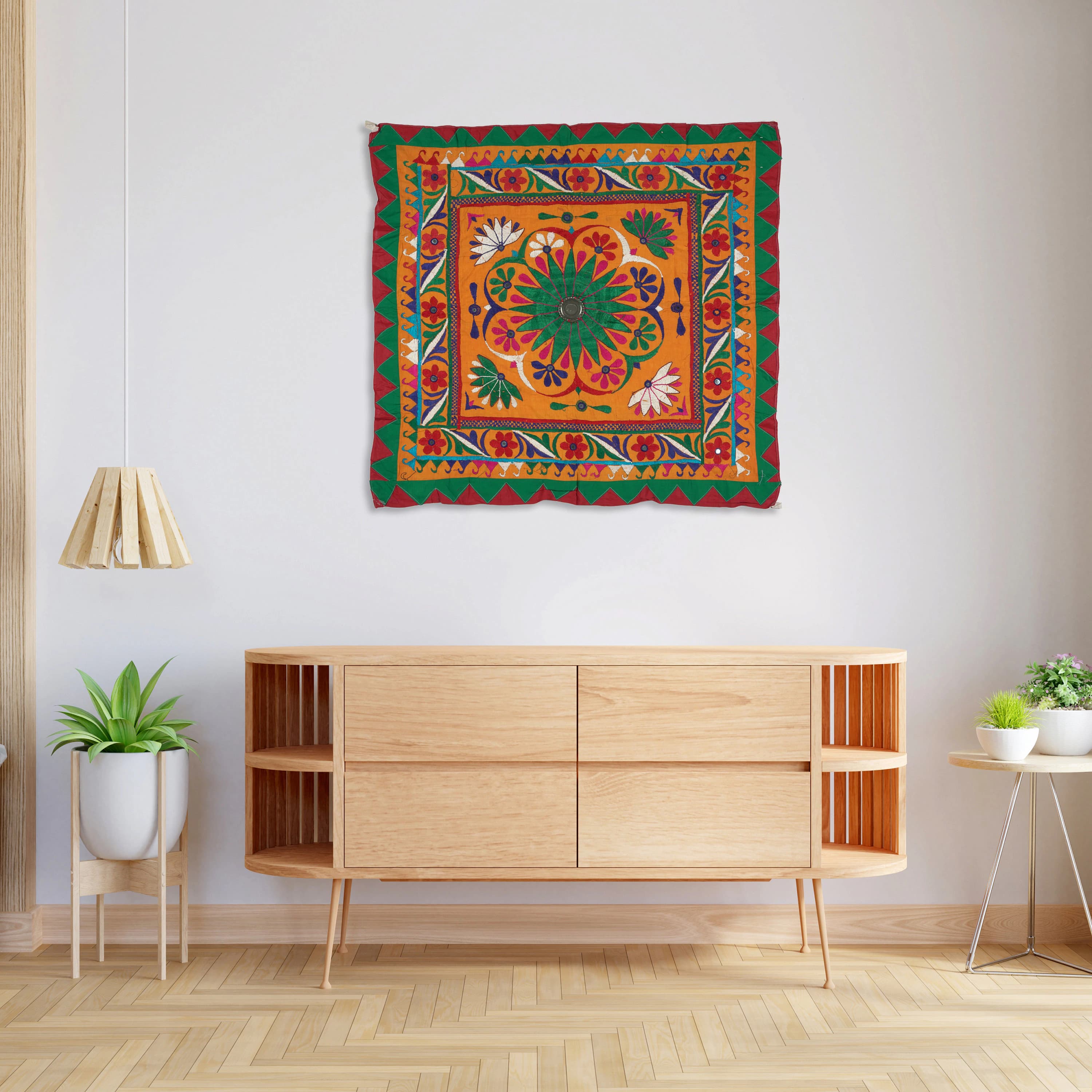 May include: A colorful wall hanging Tapestry with a square shape. The center of the hanging features a large, circular design with a green center and red, orange, yellow, and white accents. The outer border of the hanging is a dark red color with green, yellow, and white accents. The hanging is displayed on a white wall above a wooden dresser.