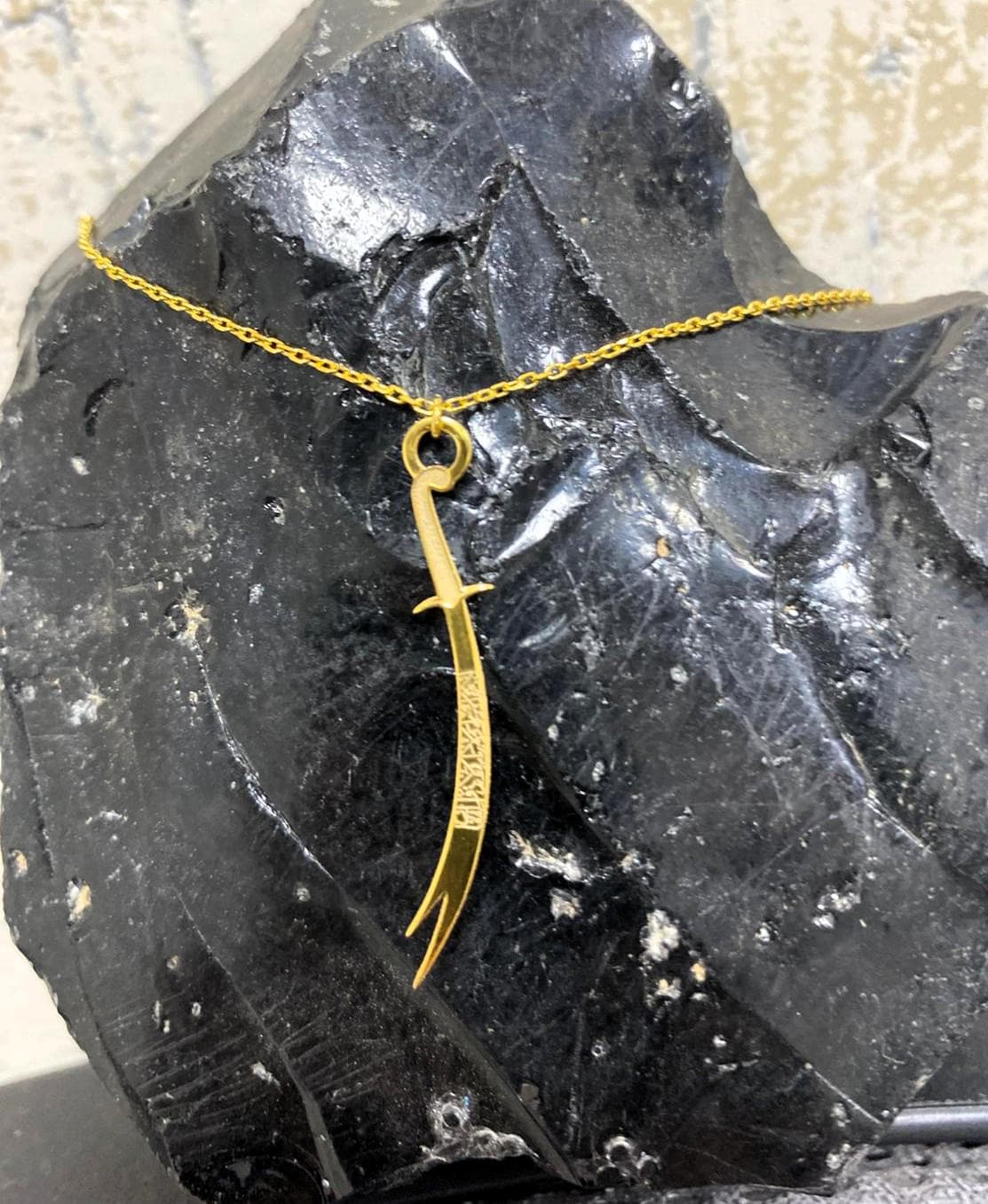 💎 PRODUCT DETAILS:
* Material: 100% Solid 925 Sterling Silver
* Finish Options: 24K Gold, Silver, Rose Gold
* Chain Length: 14” to 22” (includes charm)
* Handmade with care in our workshop A gold-colored necklace with a pendant shaped like a sword. The sword pendant has a curved blade and intricate detailing. The necklace chain is delicate and also gold-colored. The pendant is suspended against a dark, textured background.
