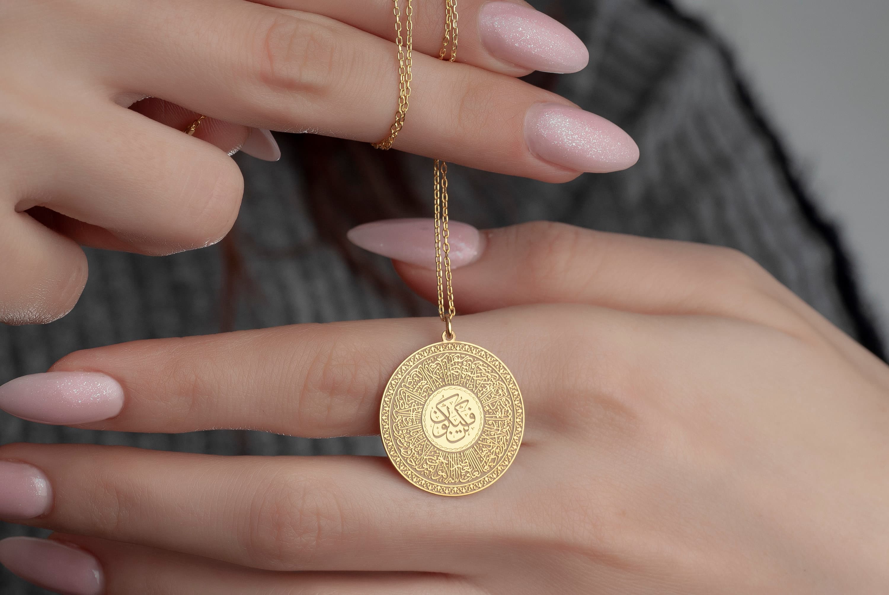 A gold necklace with a round pendant. The pendant is engraved with intricate patterns and Arabic calligraphy. The pendant is suspended from a delicate gold chain.