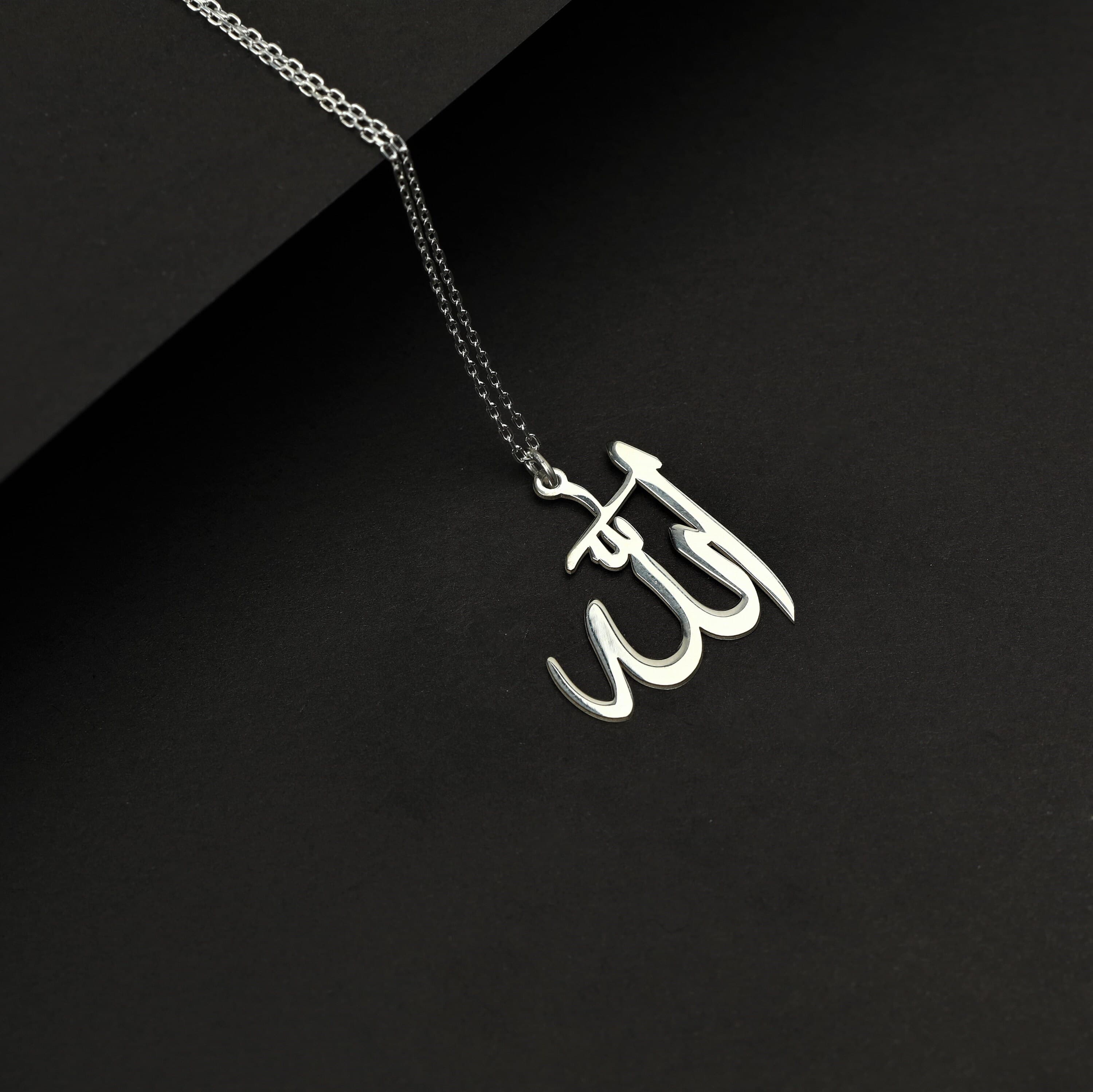 May include: A silver necklace with a pendant in the shape of the Arabic word for God, 'Allah'. The pendant is on a black background.