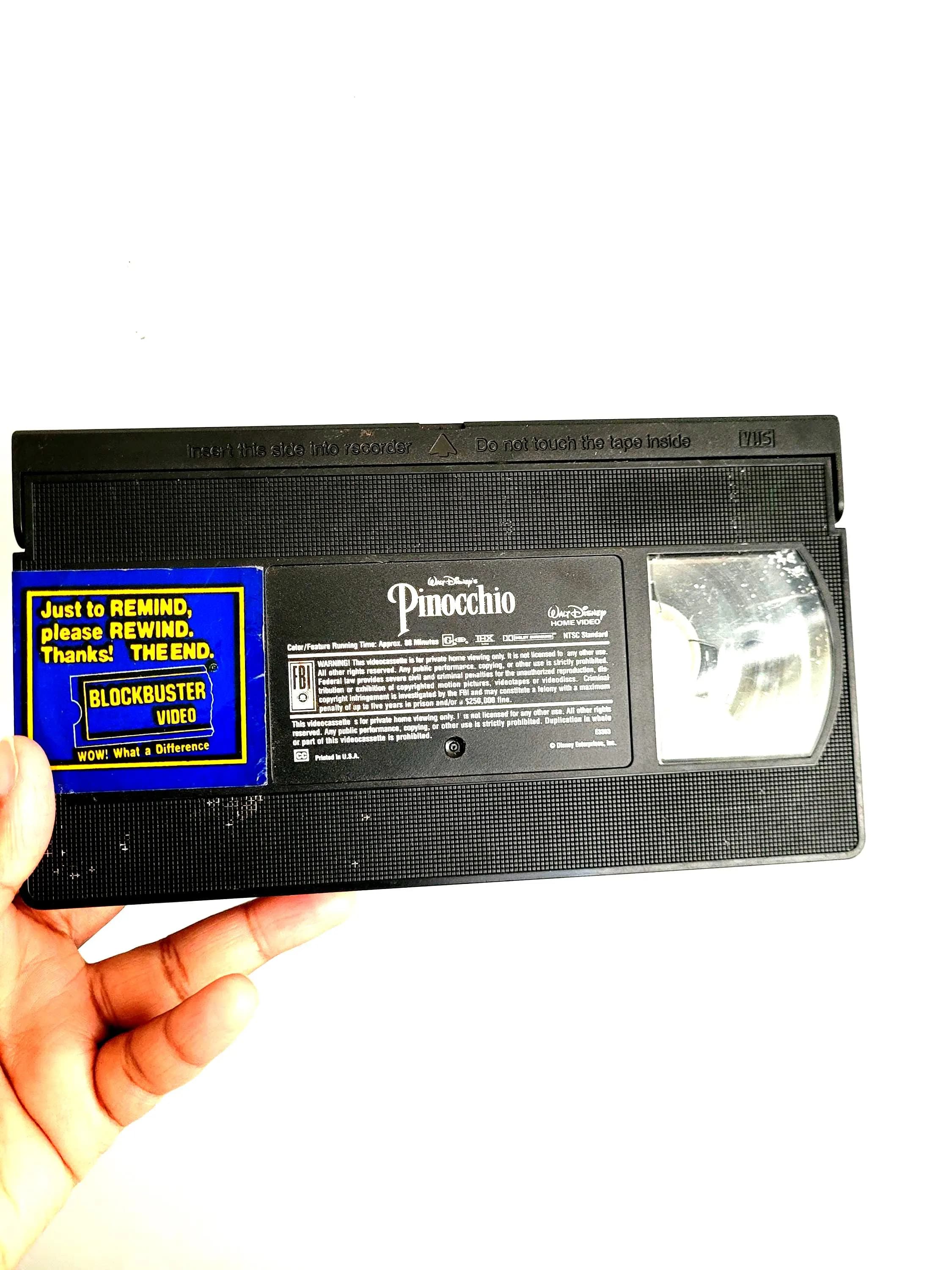 May include: A black VHS tape of Disney's Pinocchio, with a Blockbuster Video sticker. The sticker reads "Just to REMIND, please REWIND. Thanks! THE END." and "WOW! What a Difference". The tape is held against a red and patterned background.