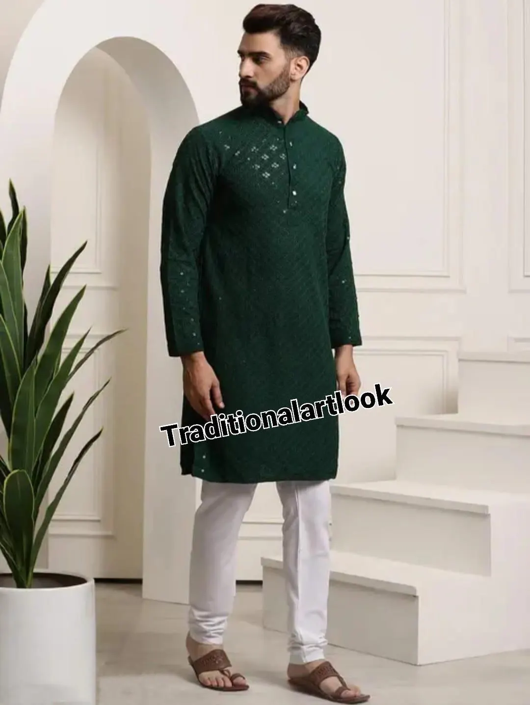 May include: A dark green tunic with a mandarin collar and intricate detailing, paired with white pants. The text "Traditionalartlook" is visible on the garment. The outfit is completed with brown sandals.