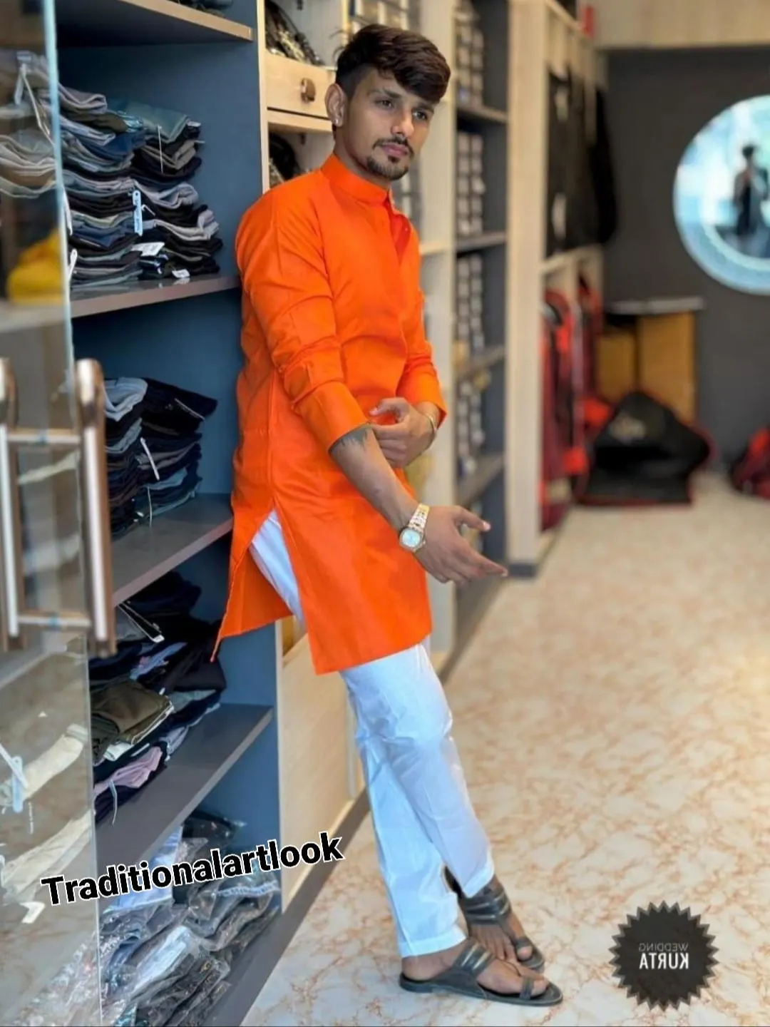 May include: An orange tunic-style shirt with long sleeves and white pants. The outfit is part of a traditional look, as indicated by the text "Traditionalartlook" on the image. The person is wearing sandals and a watch.