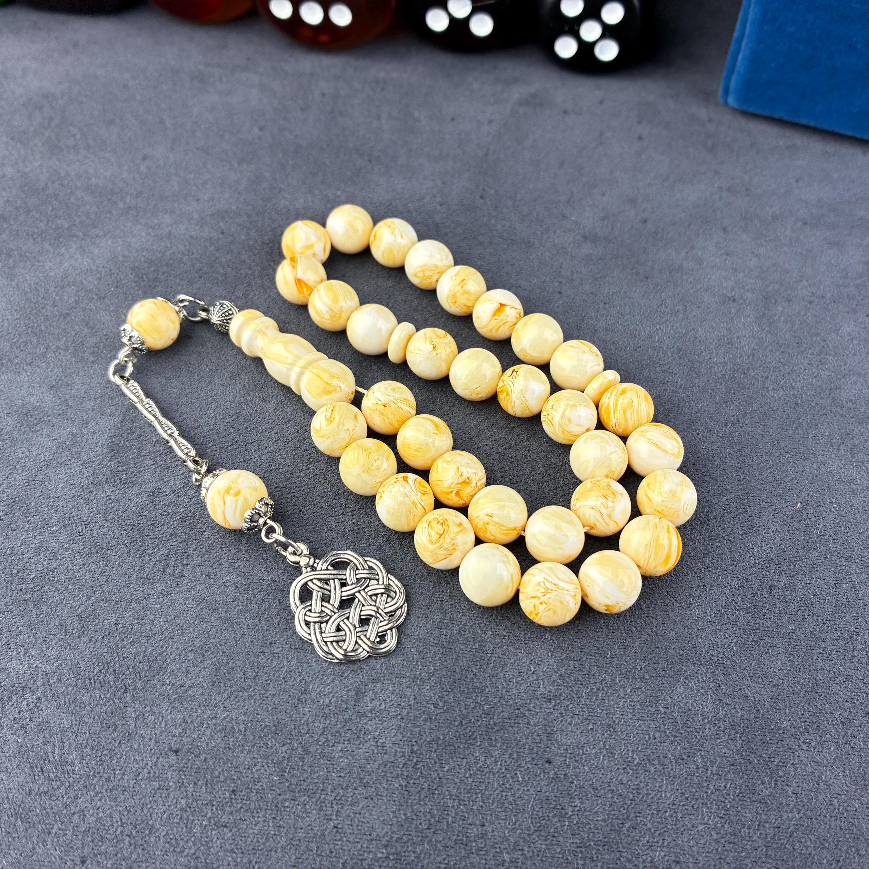 May include: A strand of prayer beads with creamy yellow beads and a silver-toned tassel. The beads have a marbled appearance. The tassel features a decorative knot design. The beads are displayed on a gray surface.