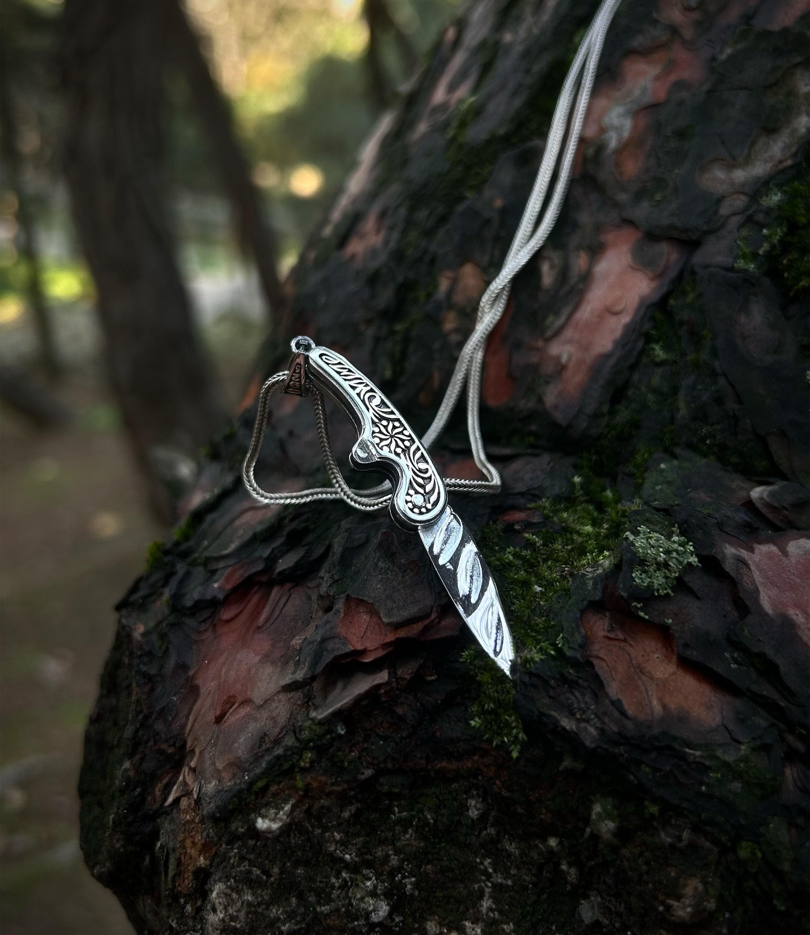 A handmade 925 sterling silver folding pocket knife necklace, shown in the open position, with an ornately engraved handle resting on mossy tree bark.