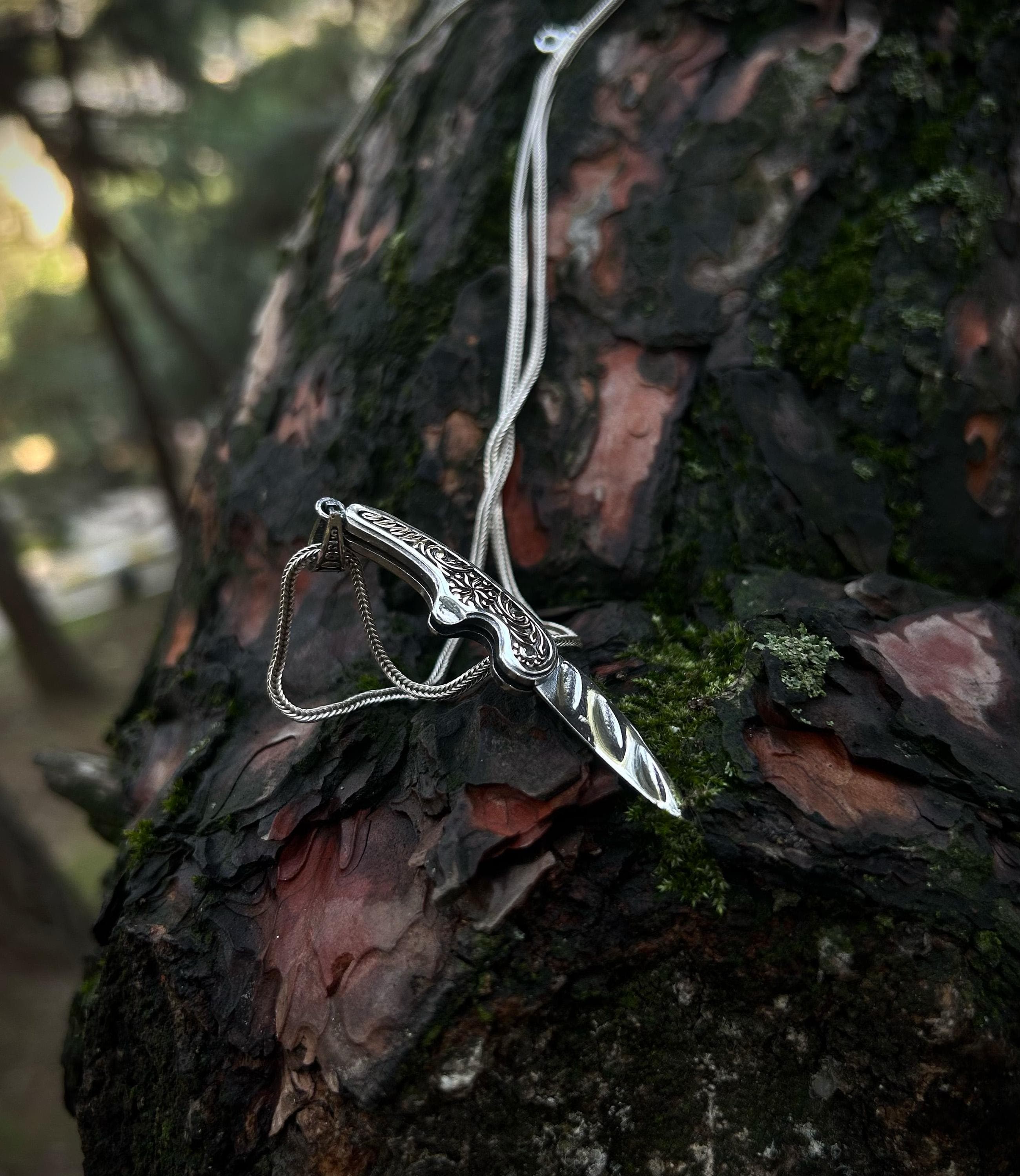 A handmade 925 sterling silver folding pocket knife necklace, shown in the open position, with an ornately engraved handle resting on mossy tree bark.