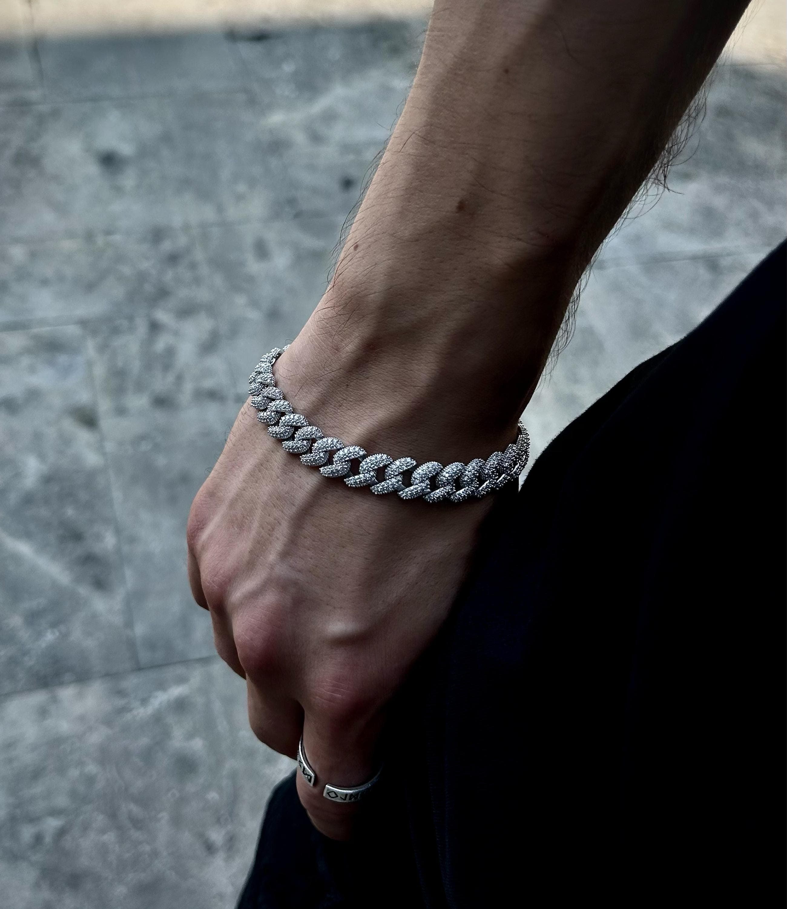 925 silver Cuban chain bracelet fully iced out with pavé stones, rhodium plated, photographed on concrete surface to reflect bold hip hop and streetwear aesthetic.