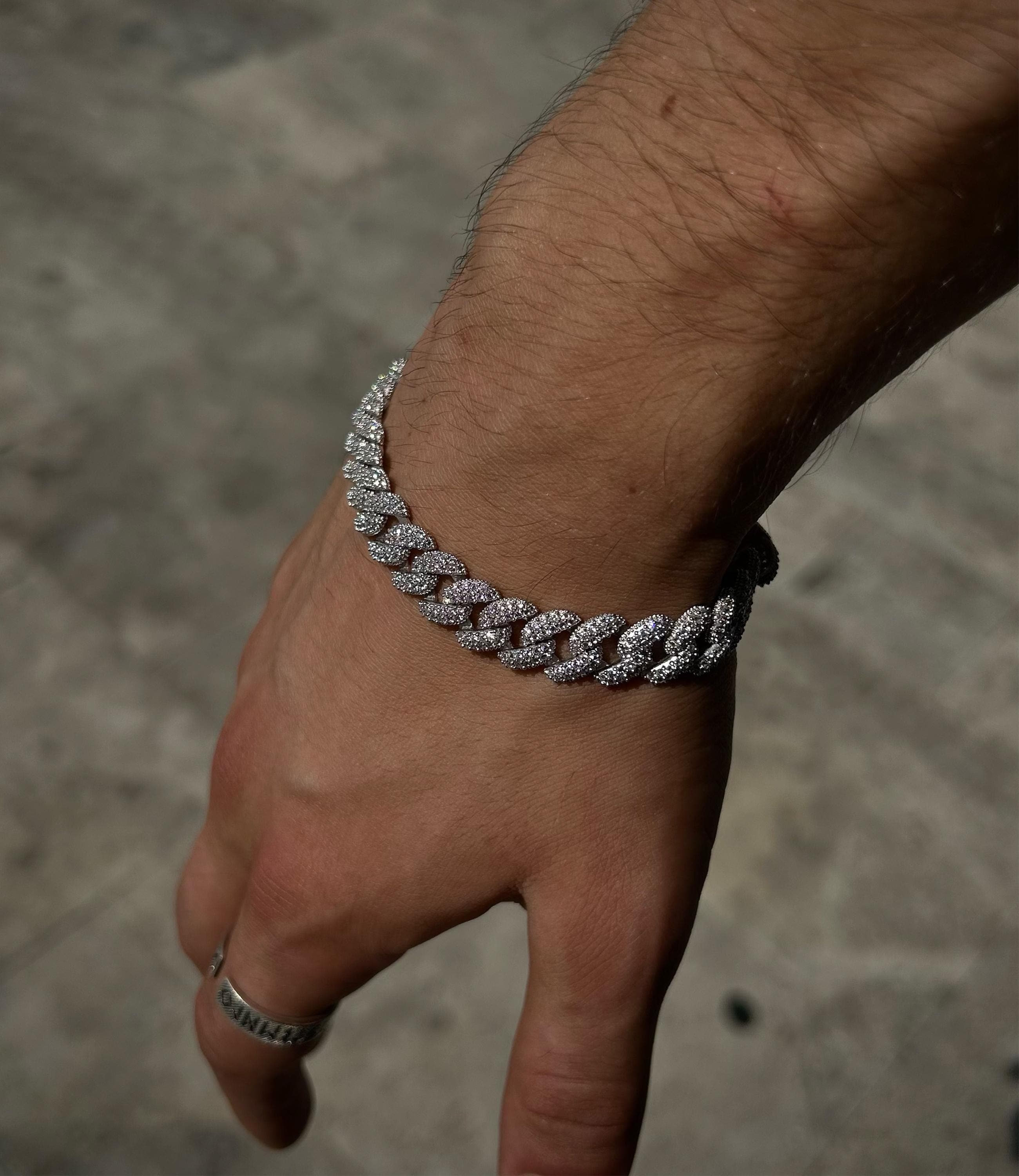 925 silver Cuban chain bracelet fully iced out with pavé stones, rhodium plated, photographed on concrete surface to reflect bold hip hop and streetwear aesthetic.