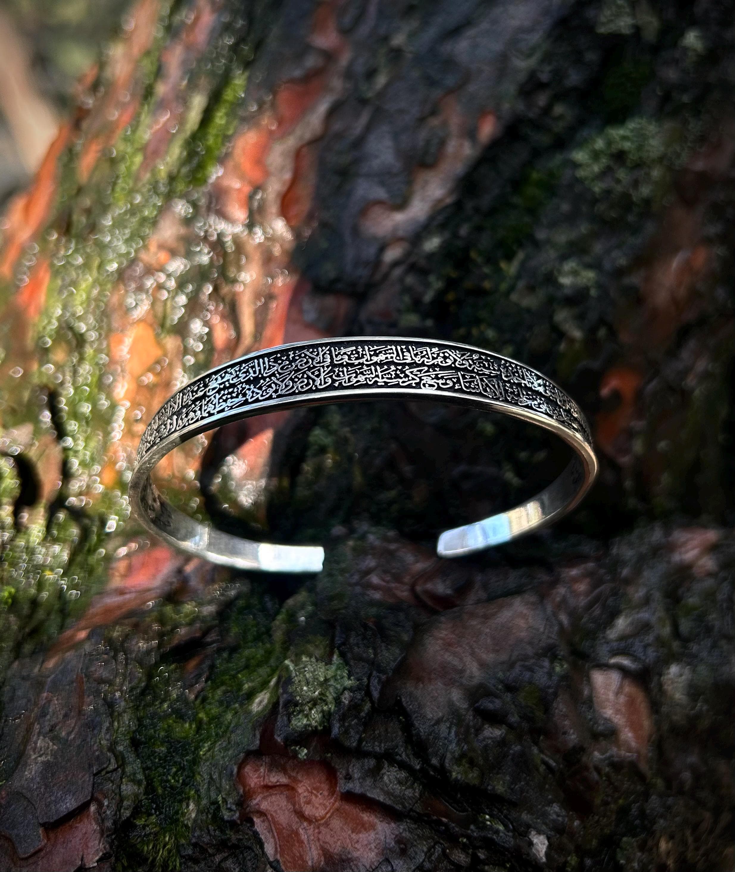 "A beautifully handcrafted 925 sterling silver cuff bracelet engraved with Ayat al-Kursi, photographed in natural light on a textured tree surface. This adjustable Islamic bangle is a meaningful spiritual jewelry piece, often worn for protection and blessings."