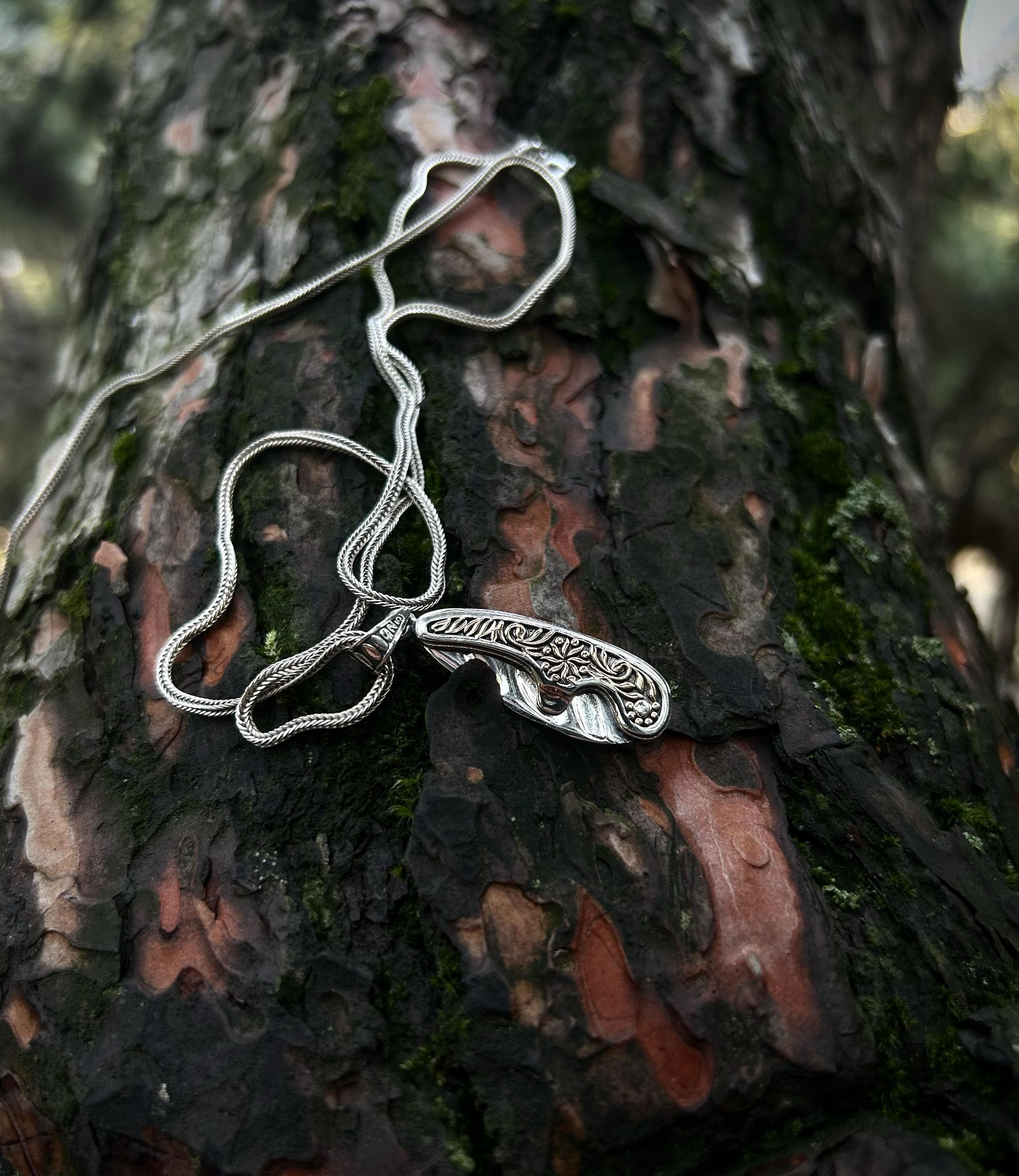 A handmade 925 sterling silver folding pocket knife necklace, shown in the open position, with an ornately engraved handle resting on mossy tree bark.