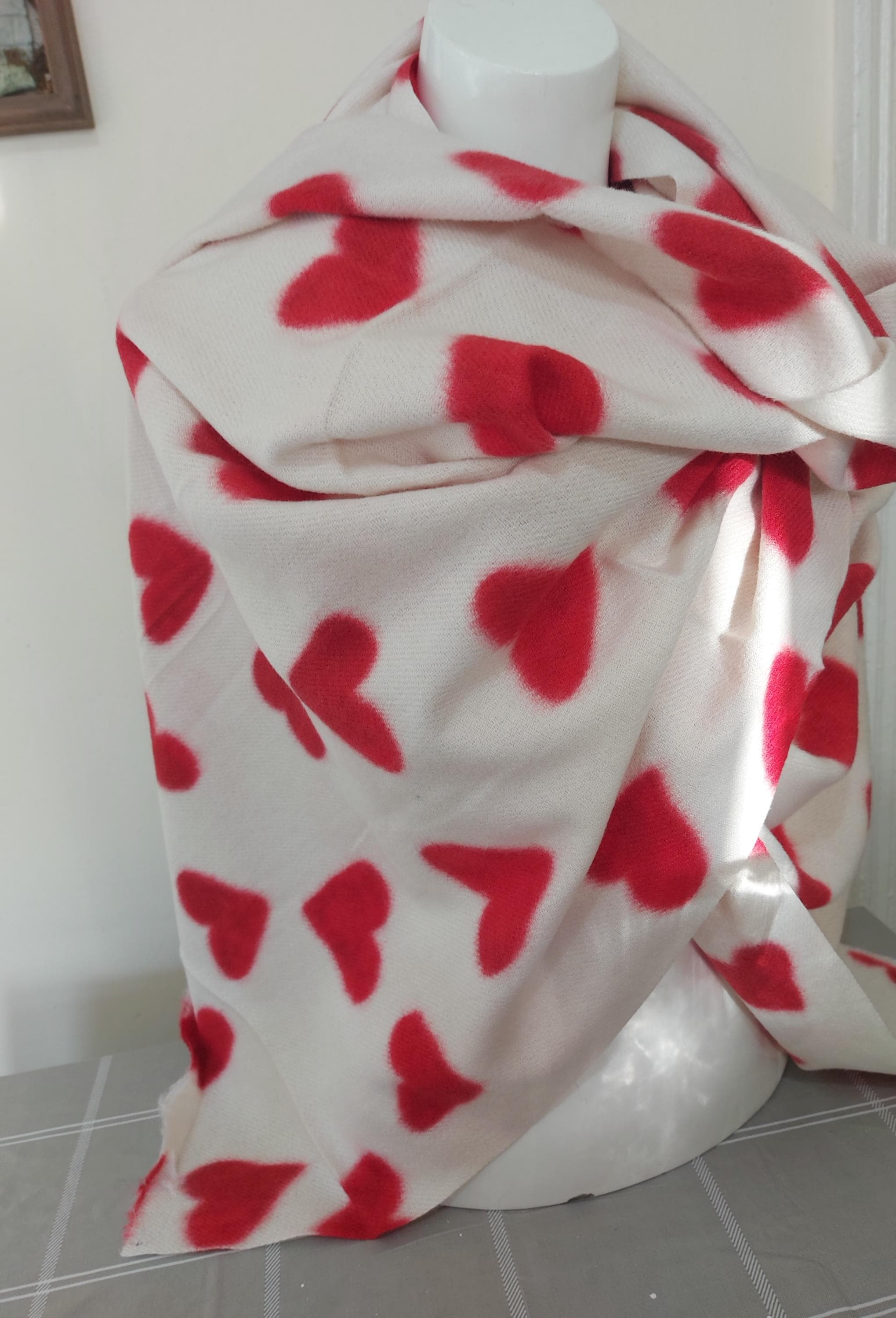 May include: A white scarf featuring a pattern of red hearts. The scarf is draped over a white form, highlighting the soft, textured fabric. The heart shapes are evenly spaced across the scarf.