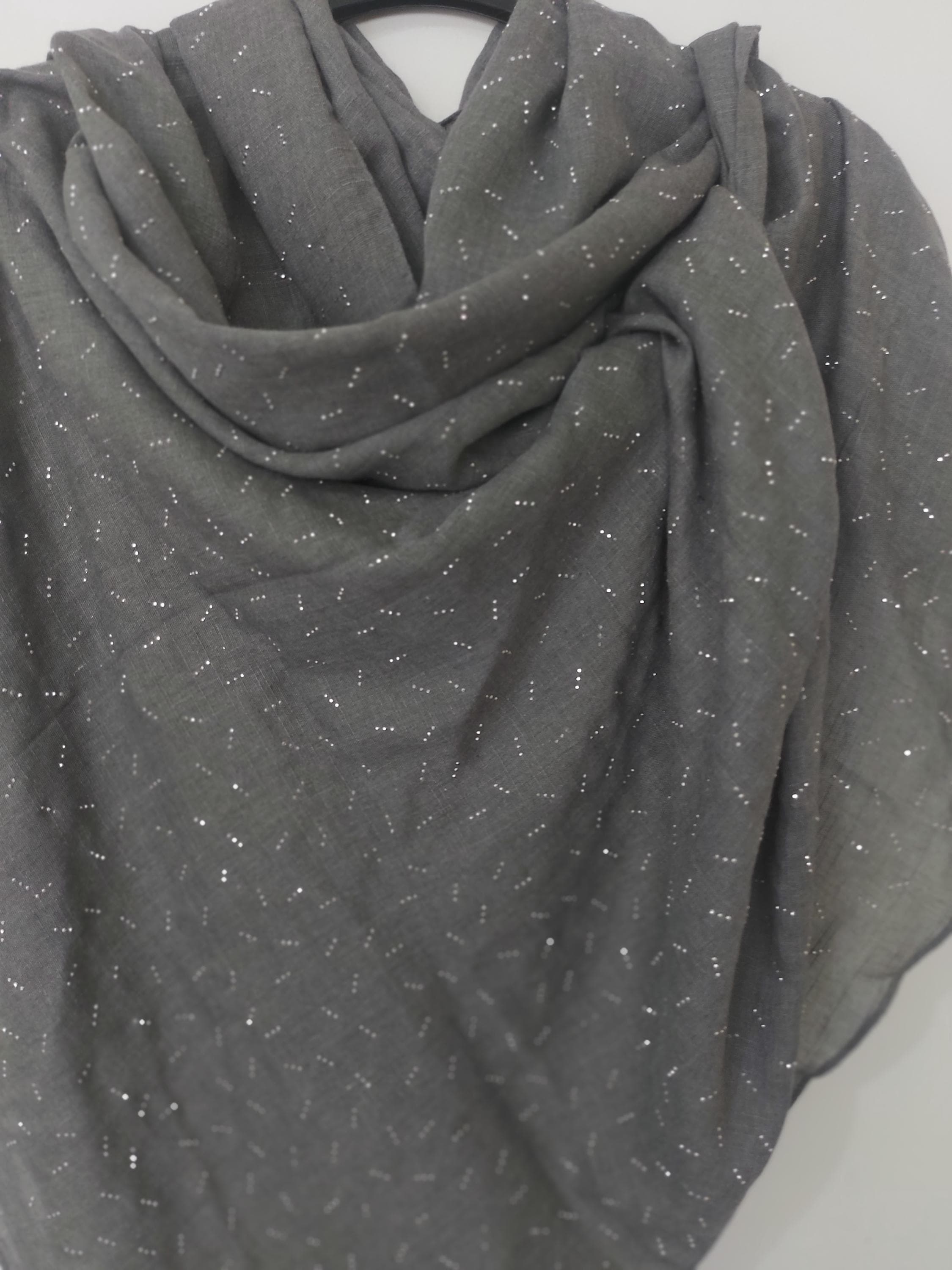 May include: A charcoal grey scarf featuring a repeating pattern of small, metallic silver dots scattered across the fabric. The scarf is draped in soft folds, showcasing the lightweight texture of the material and the shimmering effect of the silver accents.