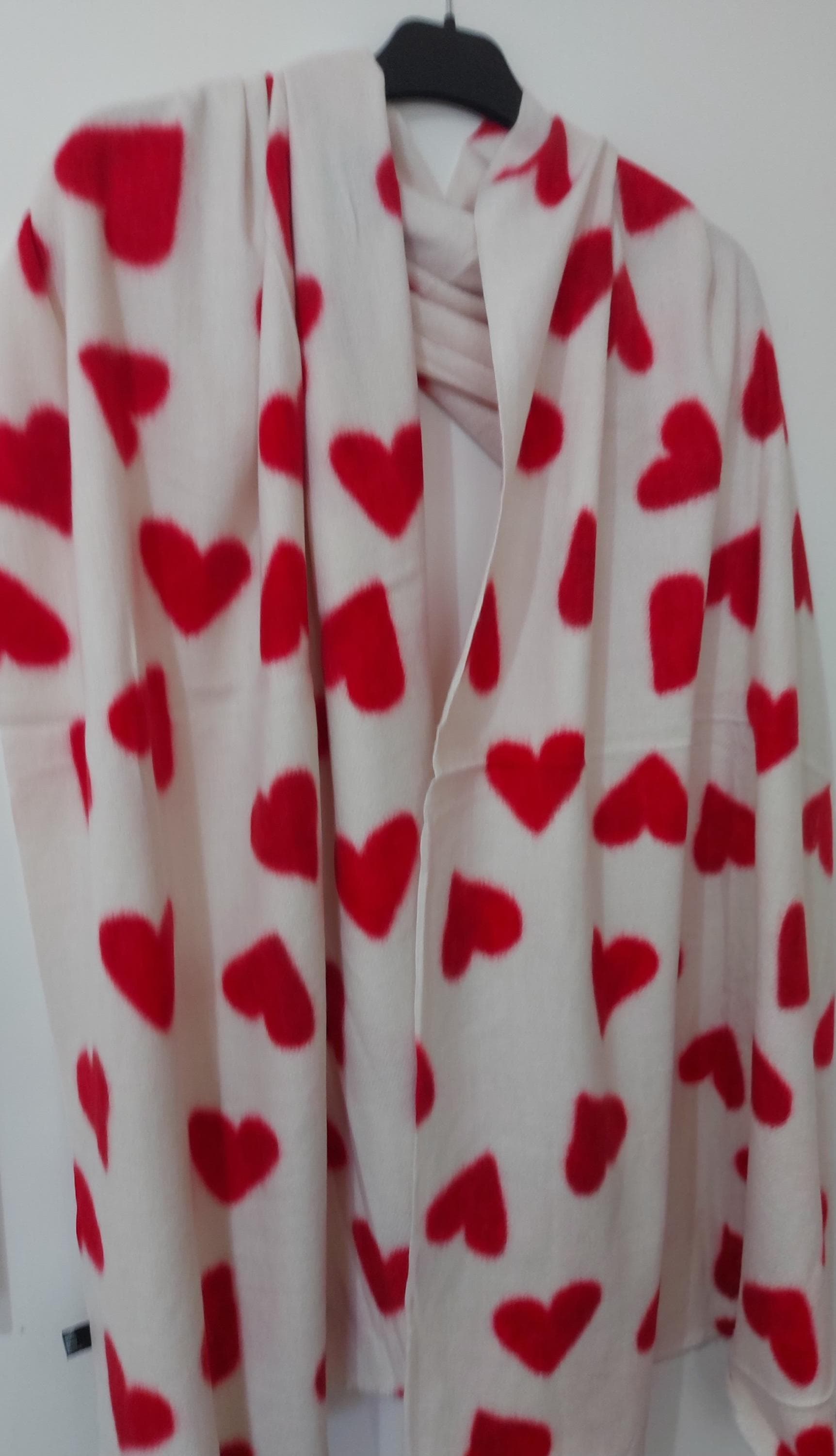 May include: A white scarf featuring a pattern of red hearts. The scarf is displayed on a hanger, highlighting the soft material and the scattered heart design. The hearts are of varying sizes and are evenly spaced across the scarf.