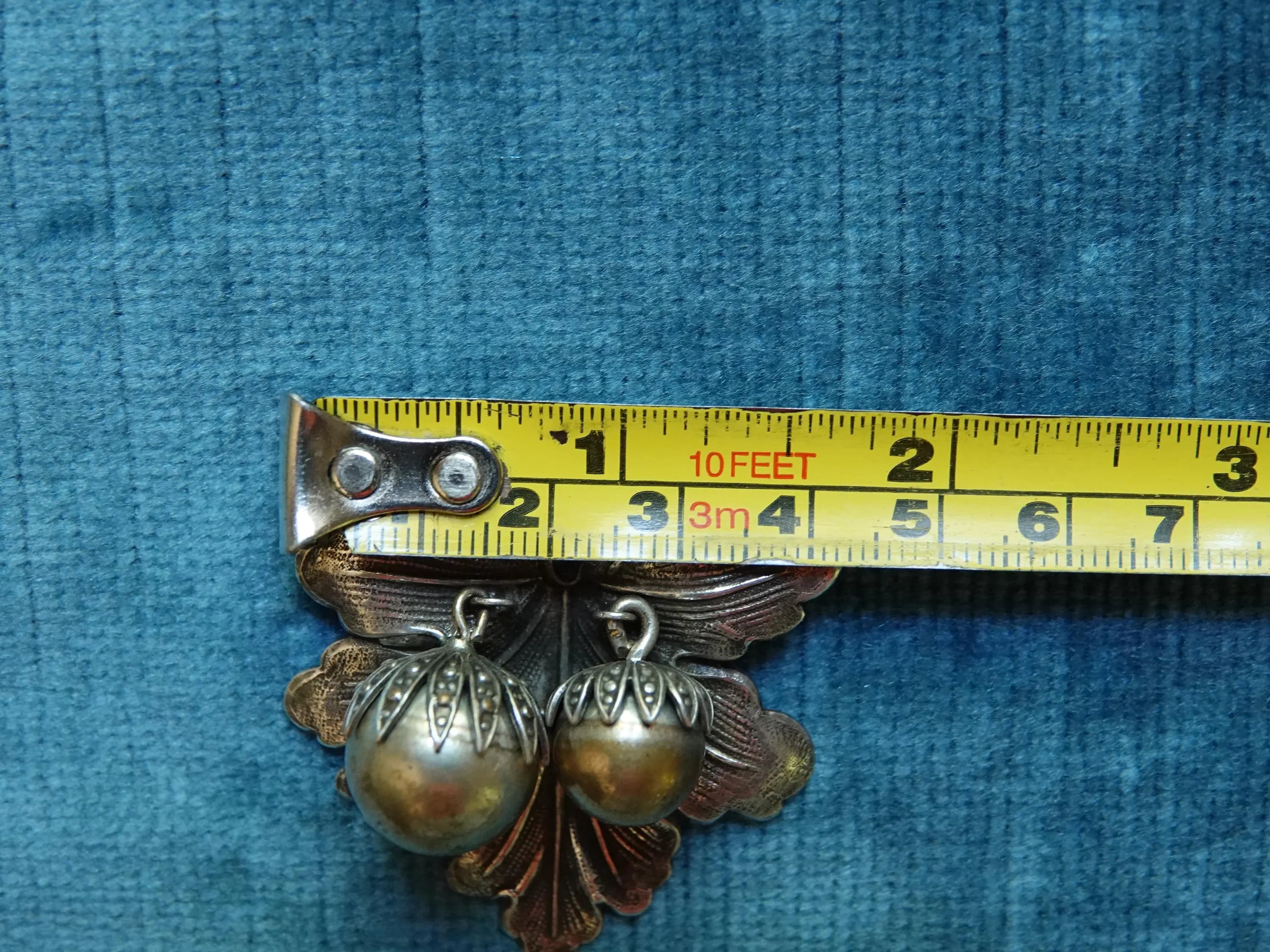 A silver brooch shaped like a leaf with two brass berry accents. shown with a tape measure