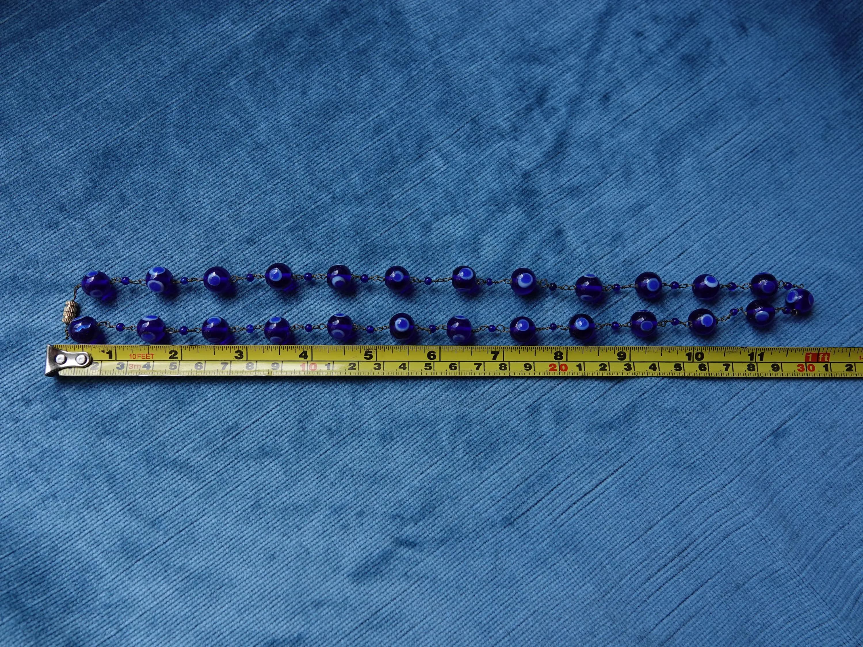Detail of beads on vintage Cobalt blue necklace with thousand eyes design. The links are brass chain with tiny cobalt blue spacers. The necklace fastens with a brass barrel fastener. Shown against a tape measure