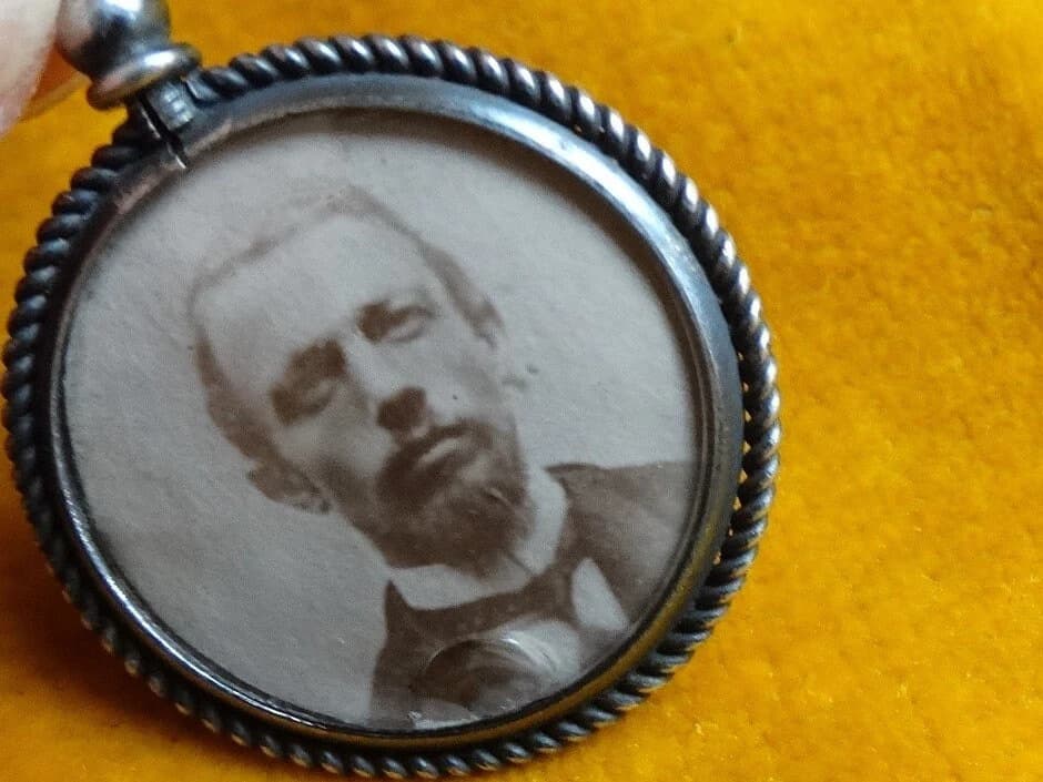 A silver locket with a braided rope design around the edge. The locket contains a seia tone photograph of a man