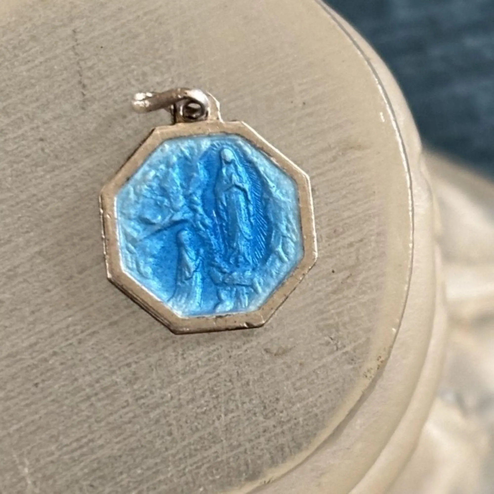 Silver and blue enamel grotto scene of The Apparition & Bernadette Soubirous at Lourdes. The tiny pendant charm has a silver octagonal border. The hanging loop has a twist in it