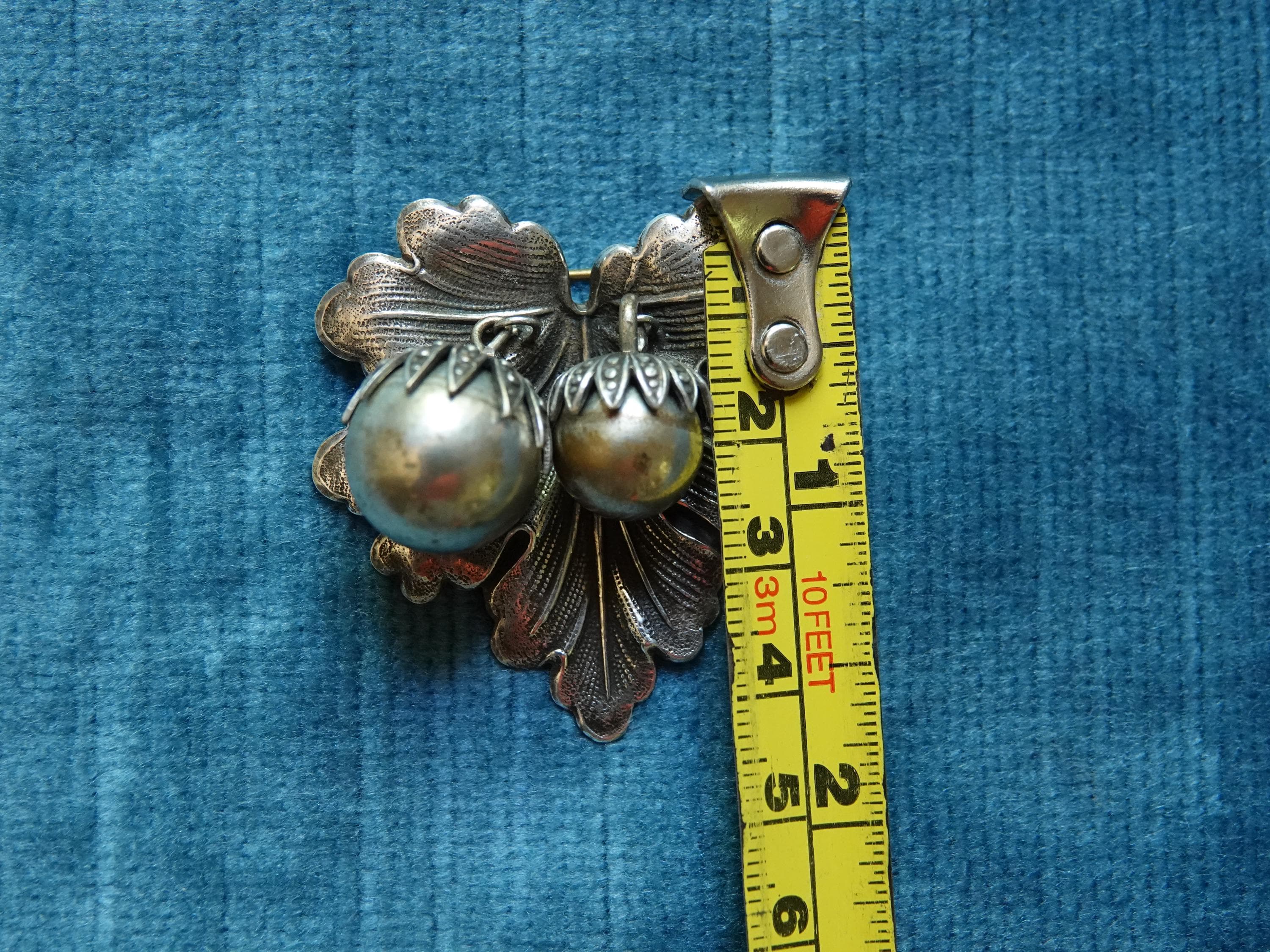 A silver brooch shaped like a leaf with two brass berry accents. shown with a tape measure