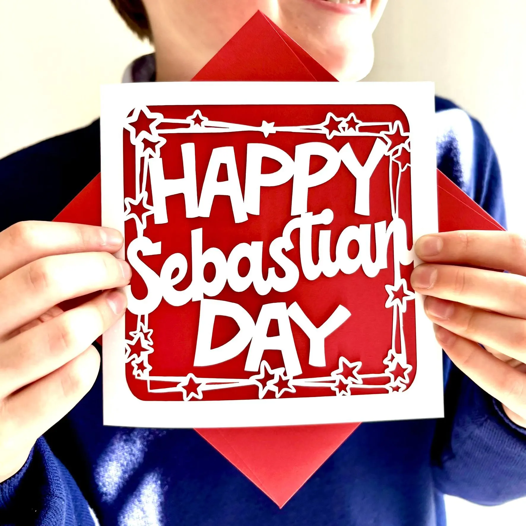 White square laser cut personalised birthday card. Featuring the words 'Happy [NAME] Day', in a bold, playful cut‑out font. The words are framed by a stylish border scattered with star outlines. The card has a red paper insert and is being held on a colour-matched envelope. The card is being held by a boy, smiling at something off camera.