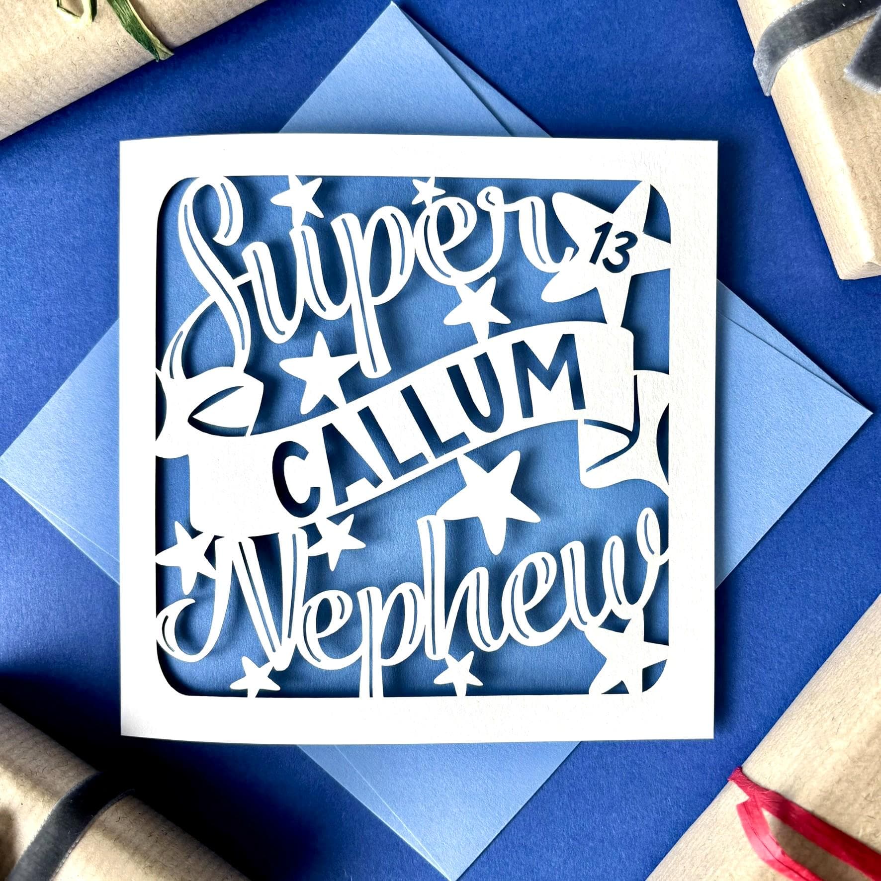 White square laser cut Nephew birthday card. Featuring the words 'Super Nephew' in a bold script font across the top and bottom of the card, personalised with a name displayed on a banner in the centre. Hand‑drawn stars surround the design. The card has a pale blue paper insert and is lying on a colour-matched envelope. The background is blue, with kraft wrapped presents visible surrounding.