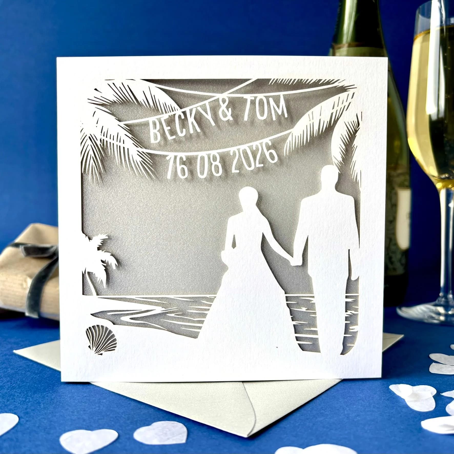 White square laser cut wedding card featuring the silhouette of a bride and groom wedding couple on a beach, surrounded by palm trees. Above the couple the card has been personalised with bunting which shows their name and the date of the wedding. The card has a silver paper insert and is standing on a colour matched envelope. It is stood against a dark blue backdrop, with heart-shaped white paper confetti, a bottle and glass of champagne and gifts visible behind.