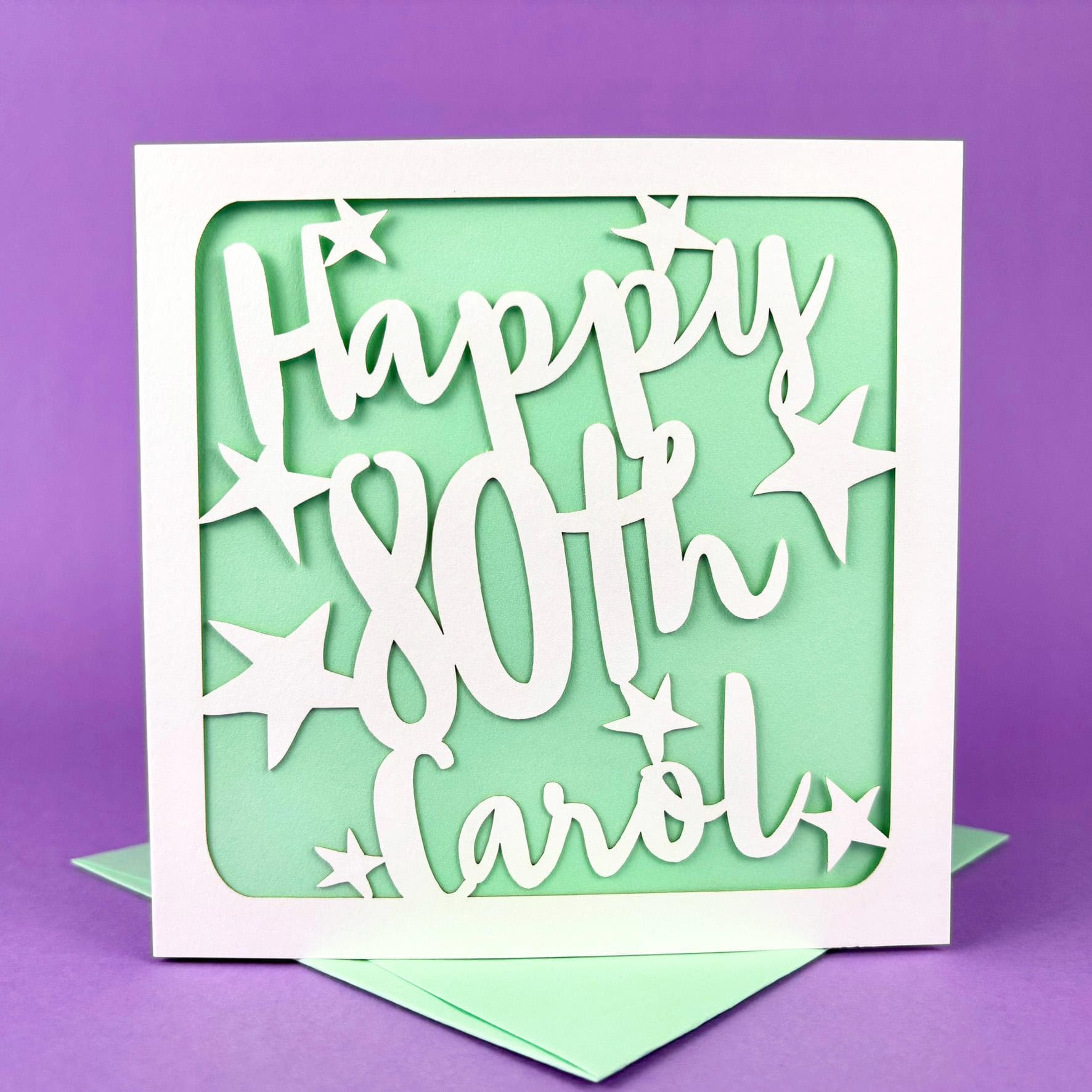 White square laser cut personalised 80th birthday card. Featuring the words 'Happy 80th' and name in bold handwritten font, surrounded by hand-drawn stars. The card has a coloured paper insert and is standing on a colour-matched envelope.